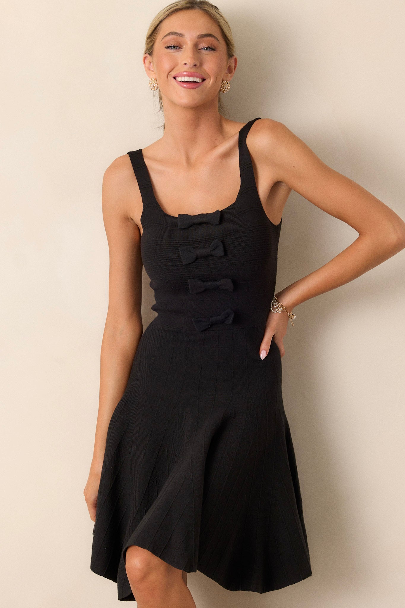 Front angled view of a black sleeveless dress featuring a square neckline, four black bow details down the bodice, a soft knit texture, and a slightly pleated skirt