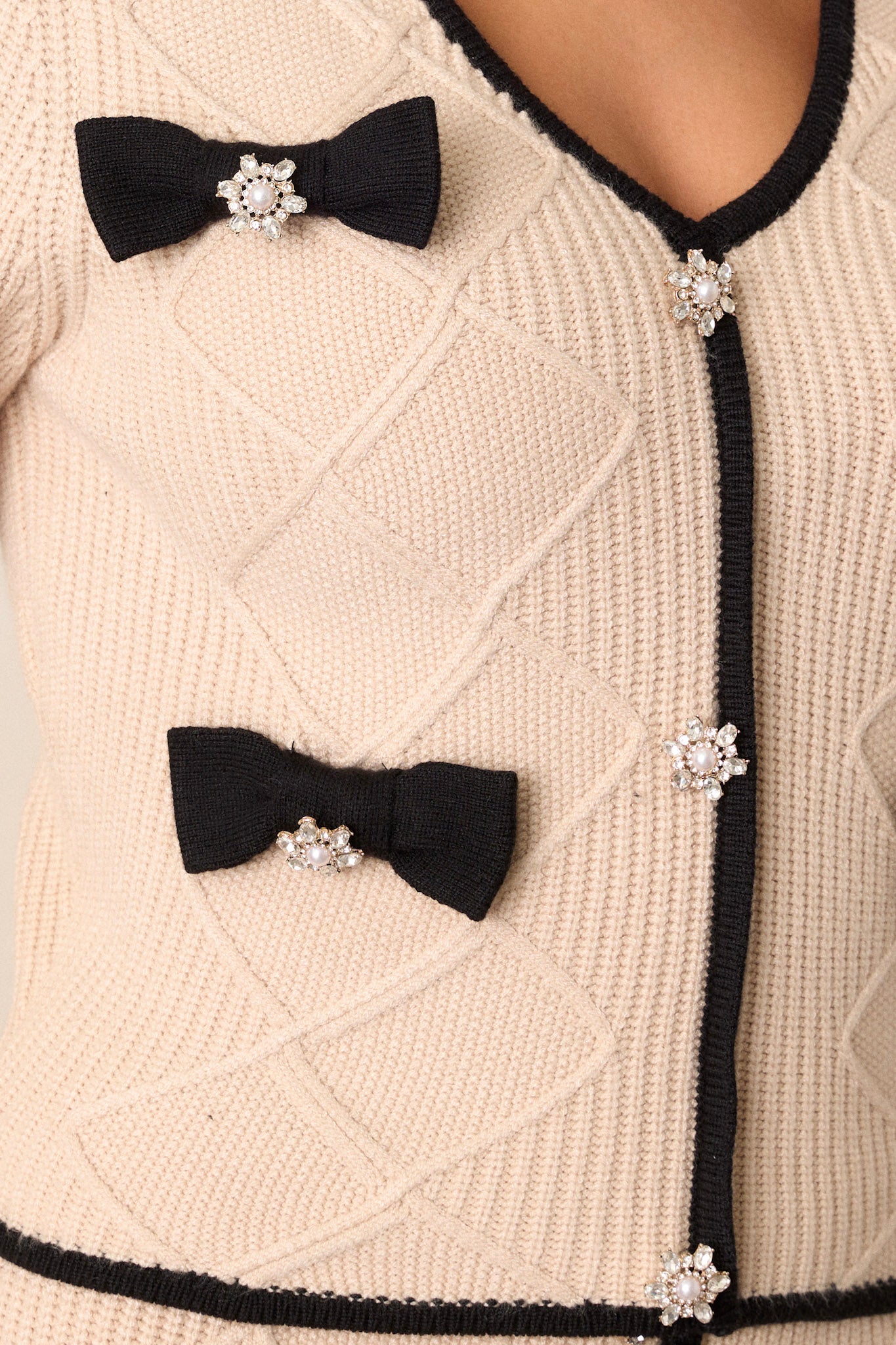 Close-up view of this beige sweater that features a v-neckline, a faux button front, black bow embellishments, faux pearl and rhinestone detailing, soft knit material, a black trim, long sleeves, and a cropped length.