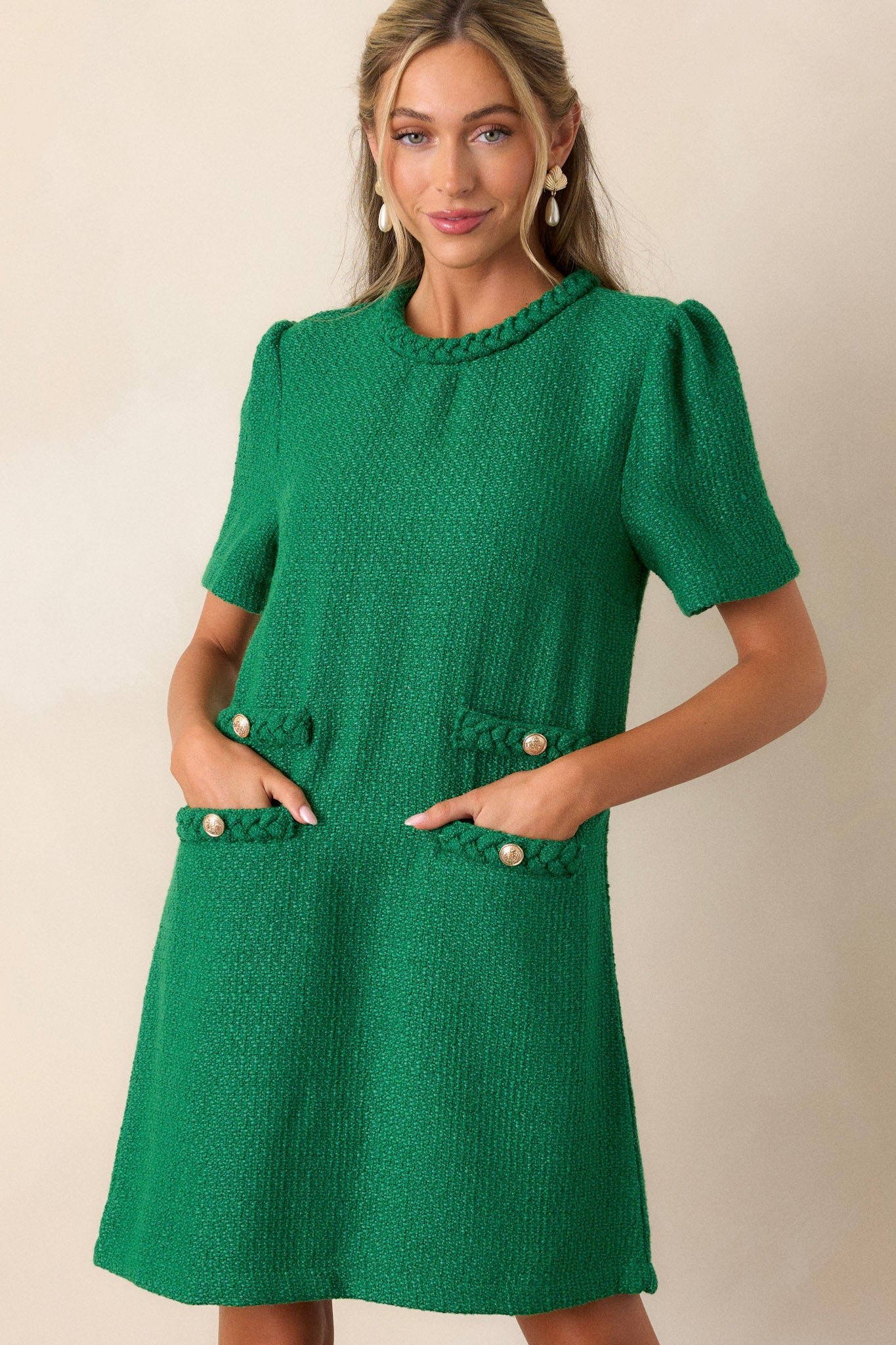 Front view of a green mini dress featuring a braided round neckline, a tweed material, functional front pockets, gold accent buttons, and a discrete back zipper