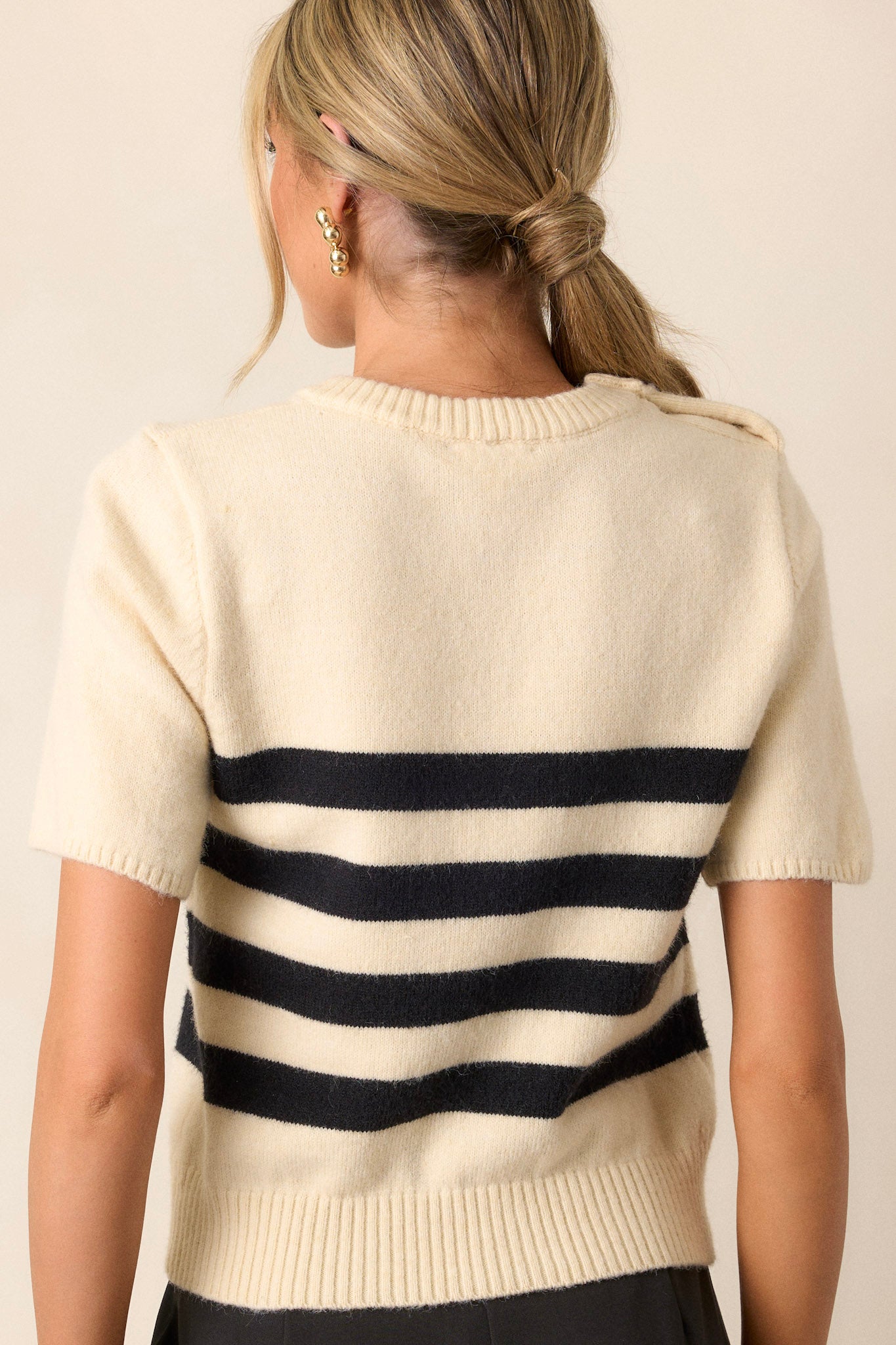 Back view of this beige sweater top featuring a ribbed crew neckline, gold accent buttons on the right shoulder, horizontal black stripes, a ribbed hemline, and short sleeves.