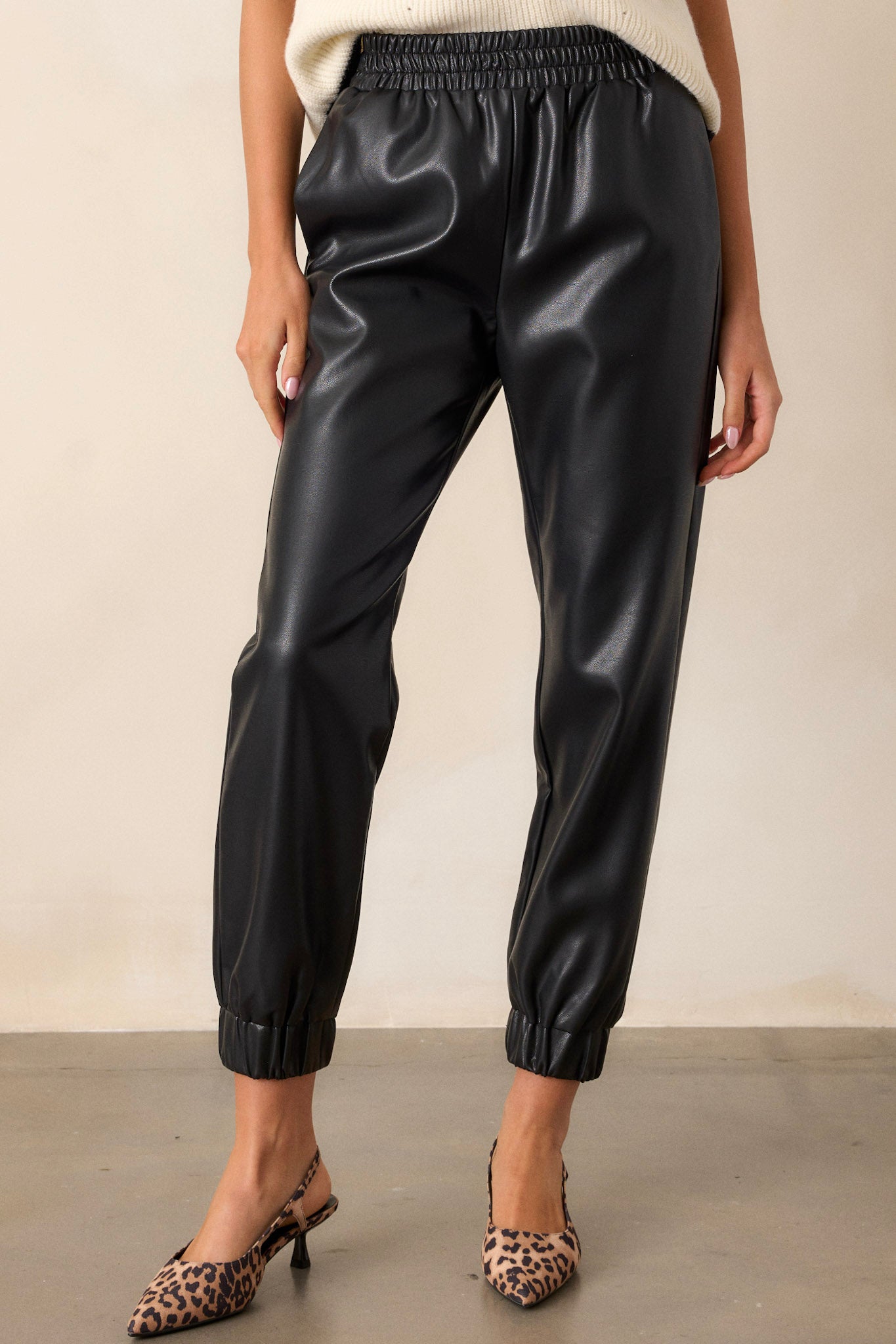 Angled Front view of black pants featuring an elastic waistband, functional hip and back side pockets, elastic cuffed ankles, and a faux leather material.