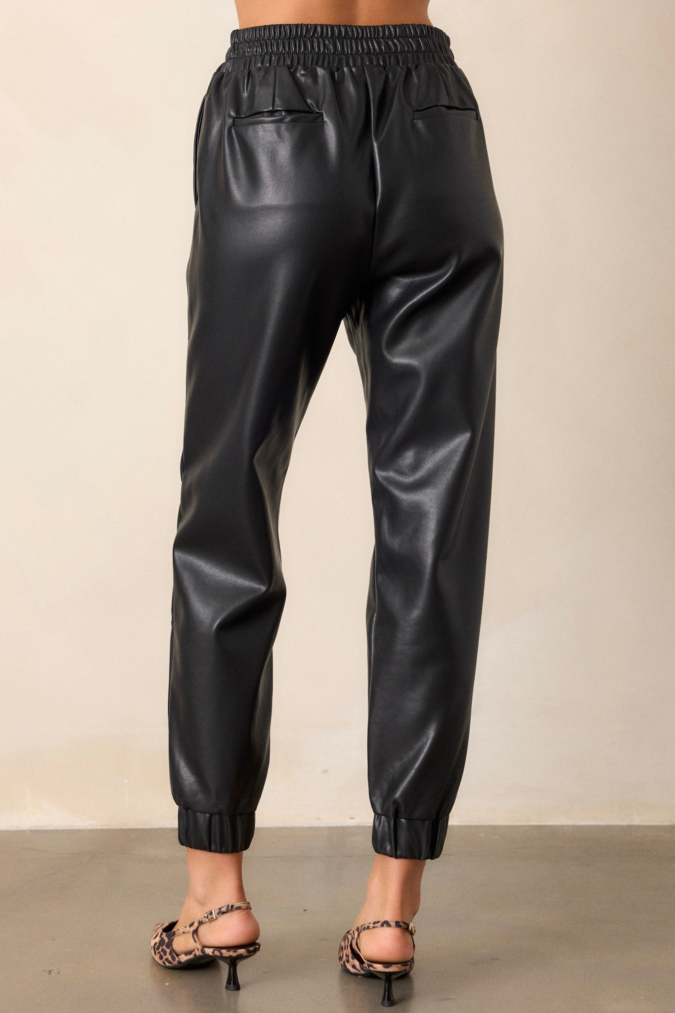 Back view of black pants featuring functional back side pockets, an elastic waistband, and elastic cuffed ankles in a faux leather material.