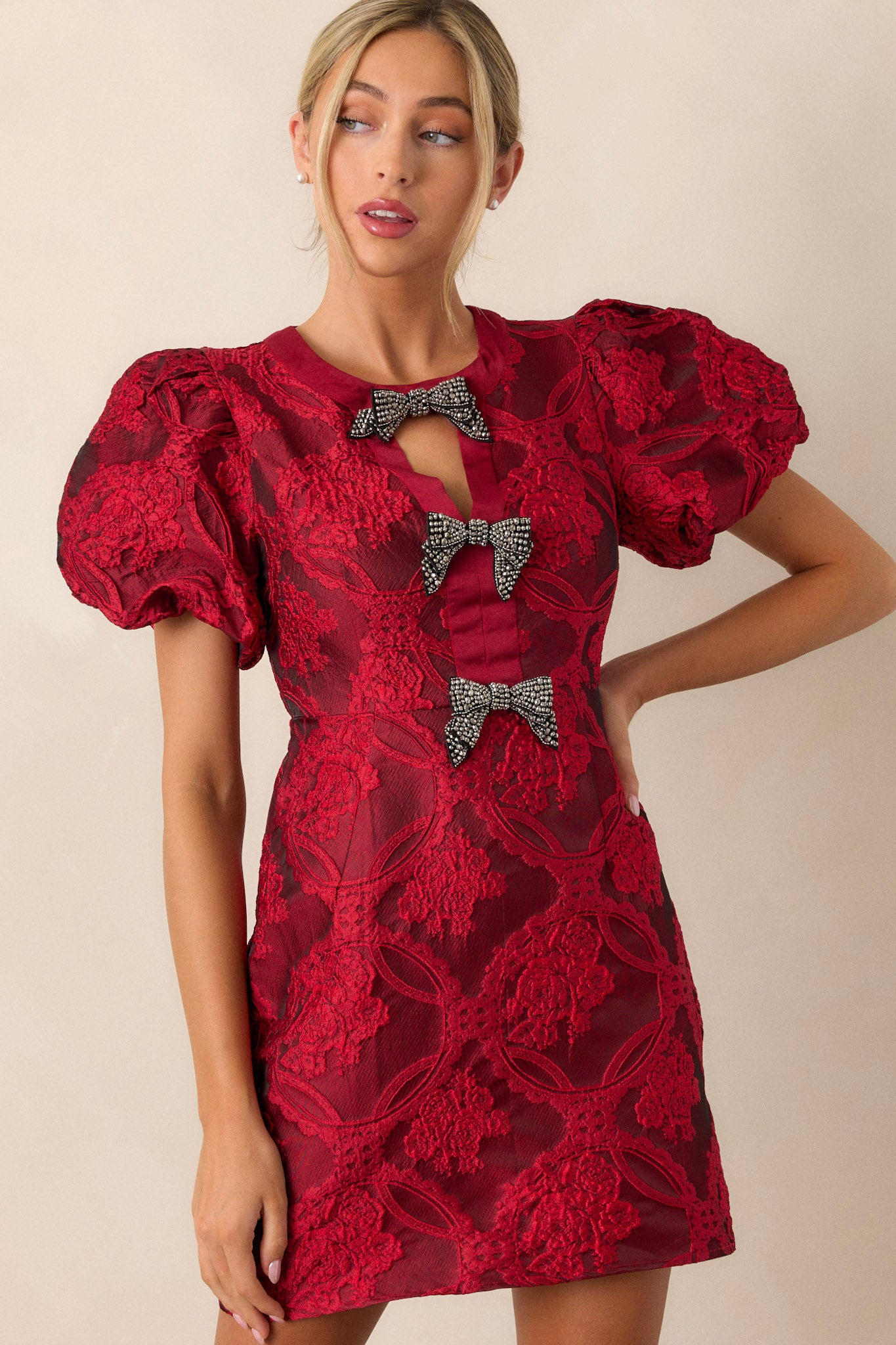 Front view of a berry red dress featuring a high neckline with a triangular cutout, three bows down the bodice, a discrete back zipper, and short elastic cuffed puff sleeves.