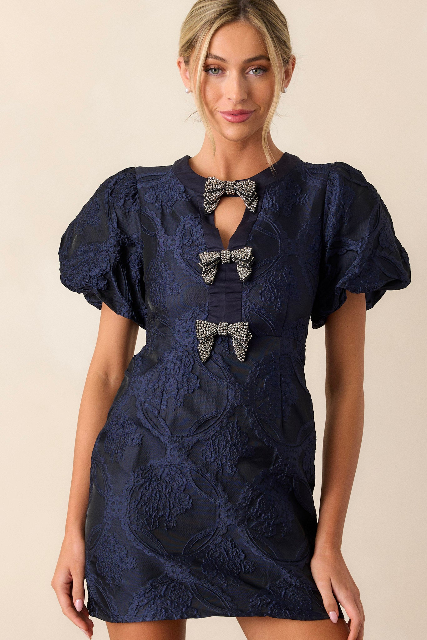 Front view of a navy dress featuring a high neckline with a triangular cutout, three bows down the bodice, and short elastic cuffed puff sleeves.