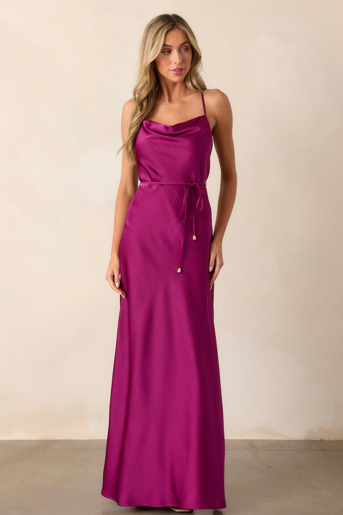 This magenta dress features a cowl neckline, adjustable straps, an adjustable self-tie belt with gold hardware, a slit reaching knee length on the left side, and a silky satin material throughout.