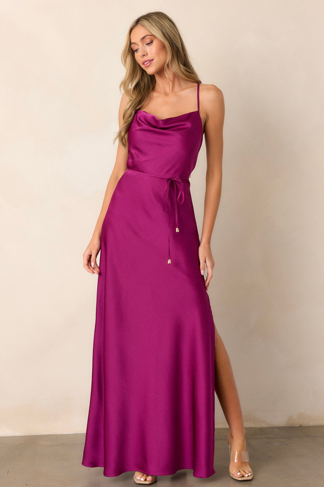 Action shot of the magenta dress displaying the fluidity and movement of the satin fabric, highlighting the cowl neckline, adjustable straps, the self-tie belt with gold hardware, and the slit on the left side.