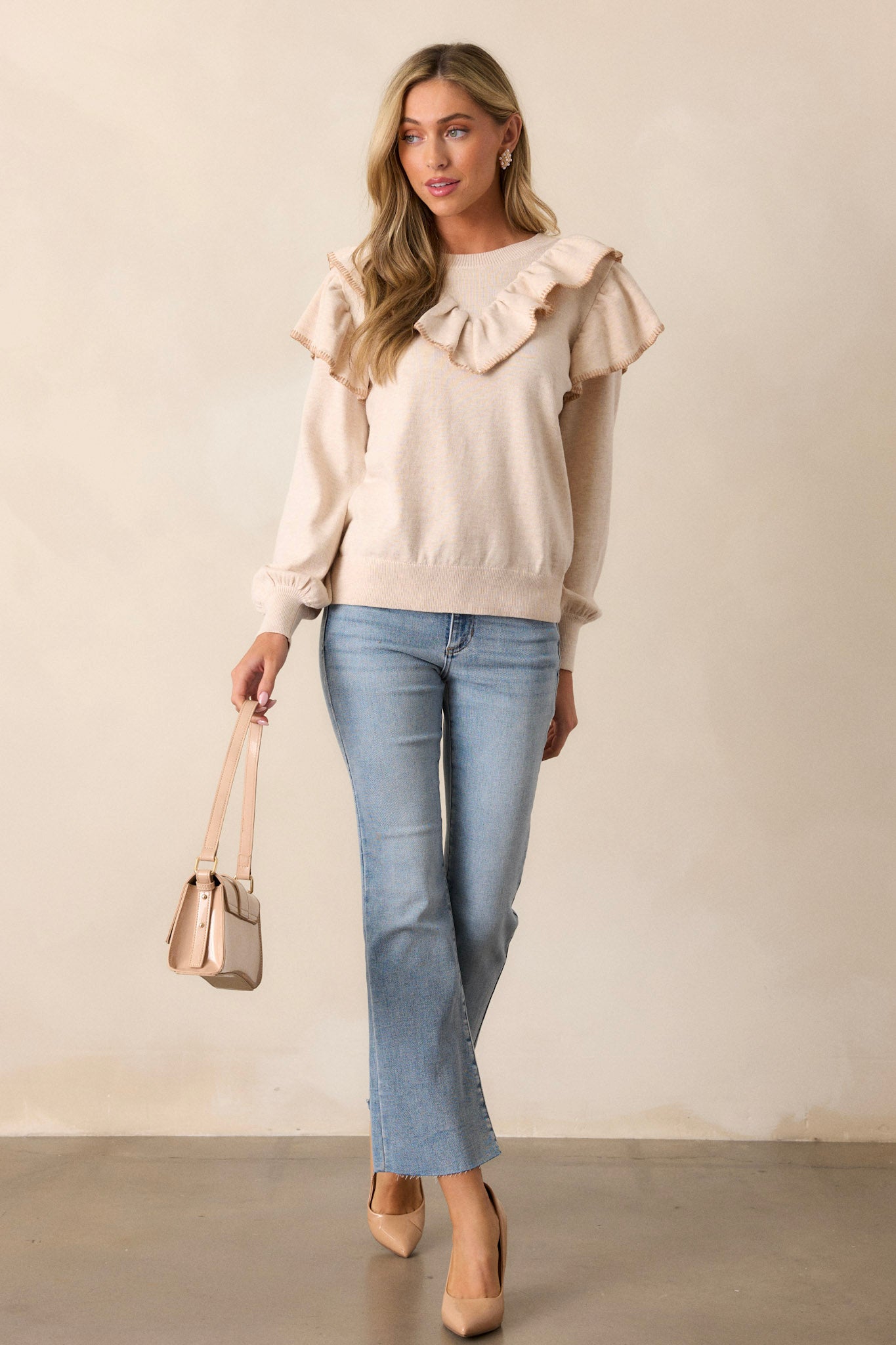 Full-length view of a model wearing the beige top, emphasizing the high neckline, ruffle detail across the shoulders and chest, ribbed hemline, and cuffed long sleeves with ruffle detailing.