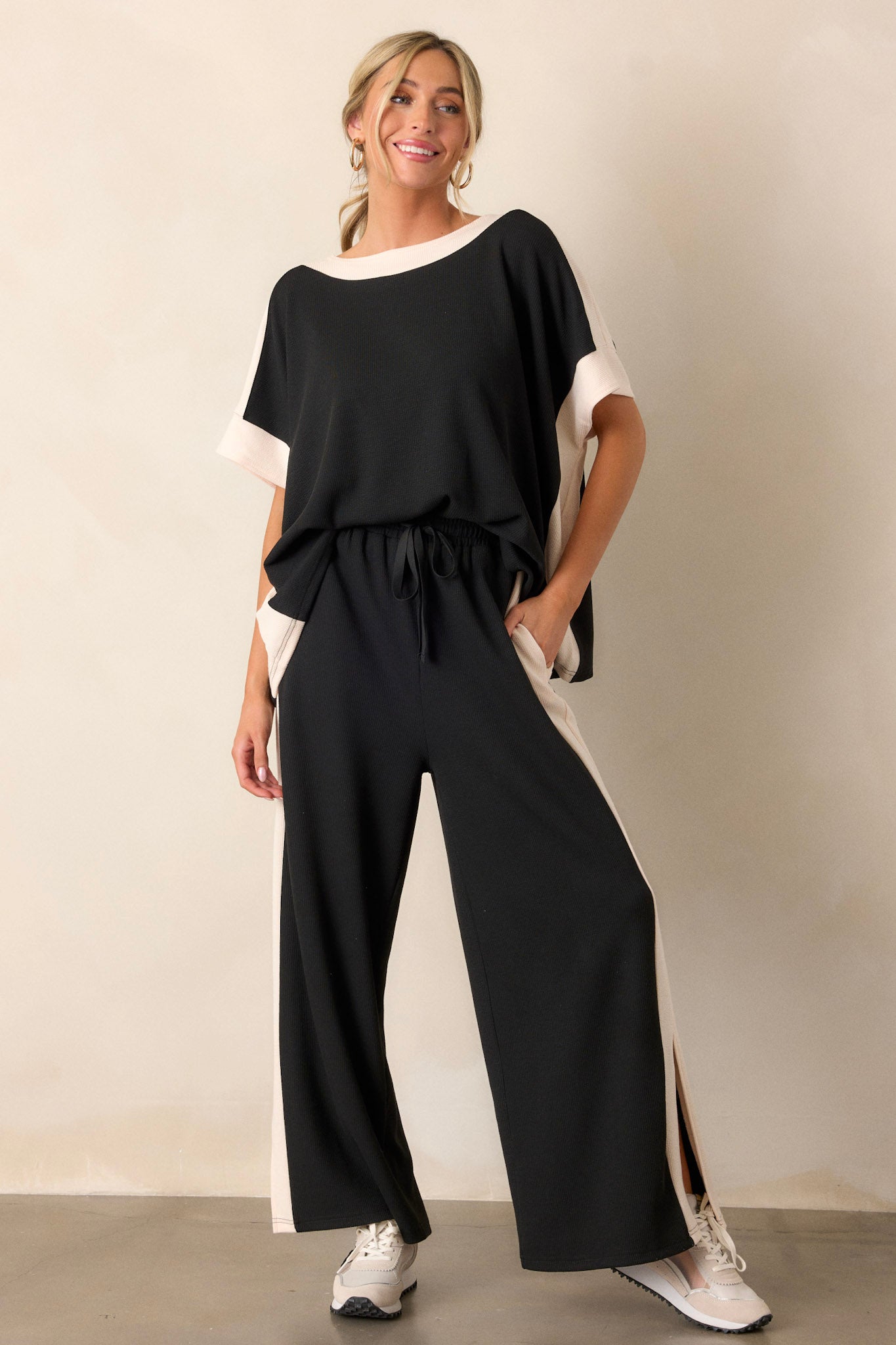 Full length view of the black and ivory pants featuring an elastic waistband, a high waisted design, a waist drawstring, soft ribbed texture, functional hip pockets, white trim down the sides, and high slits in the legs.