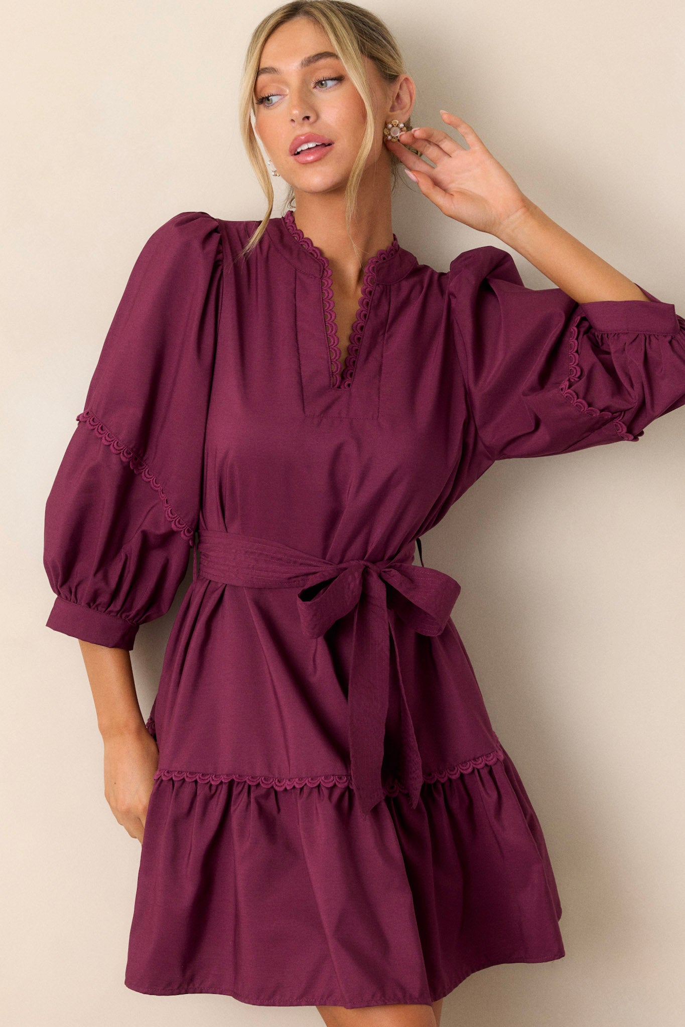 Full length view of a plum mini dress featuring a split neckline with scalloped detailing, a self-tie drawstring belt, functional hip pockets, a tiered design, and tiered bishop sleeves with scalloped detailing