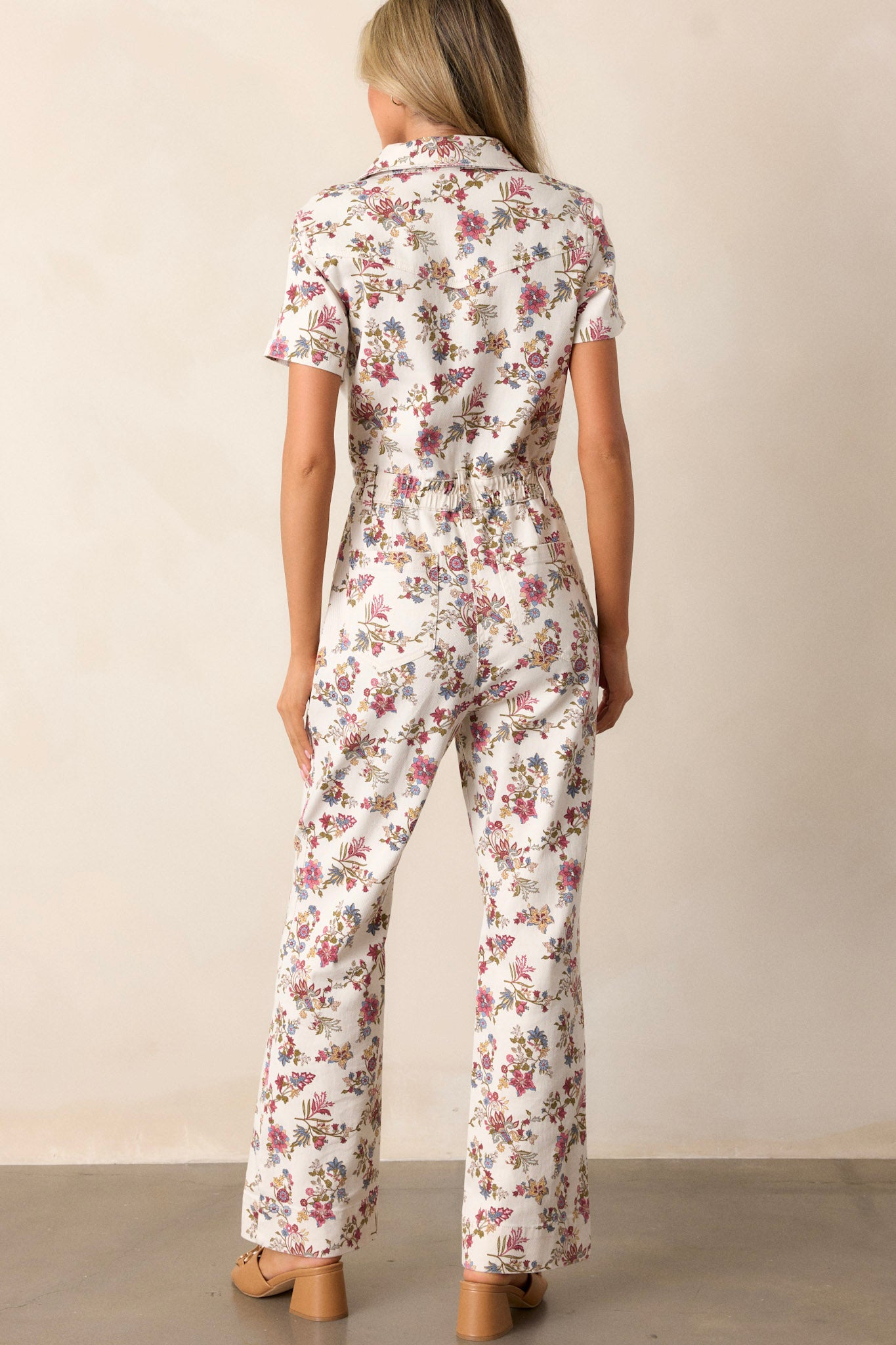 Back view of this ivory denim jumpsuit that features four functional pockets, a functional zipper, a collared neckline, a button-front bust, and a floral denim fabric.