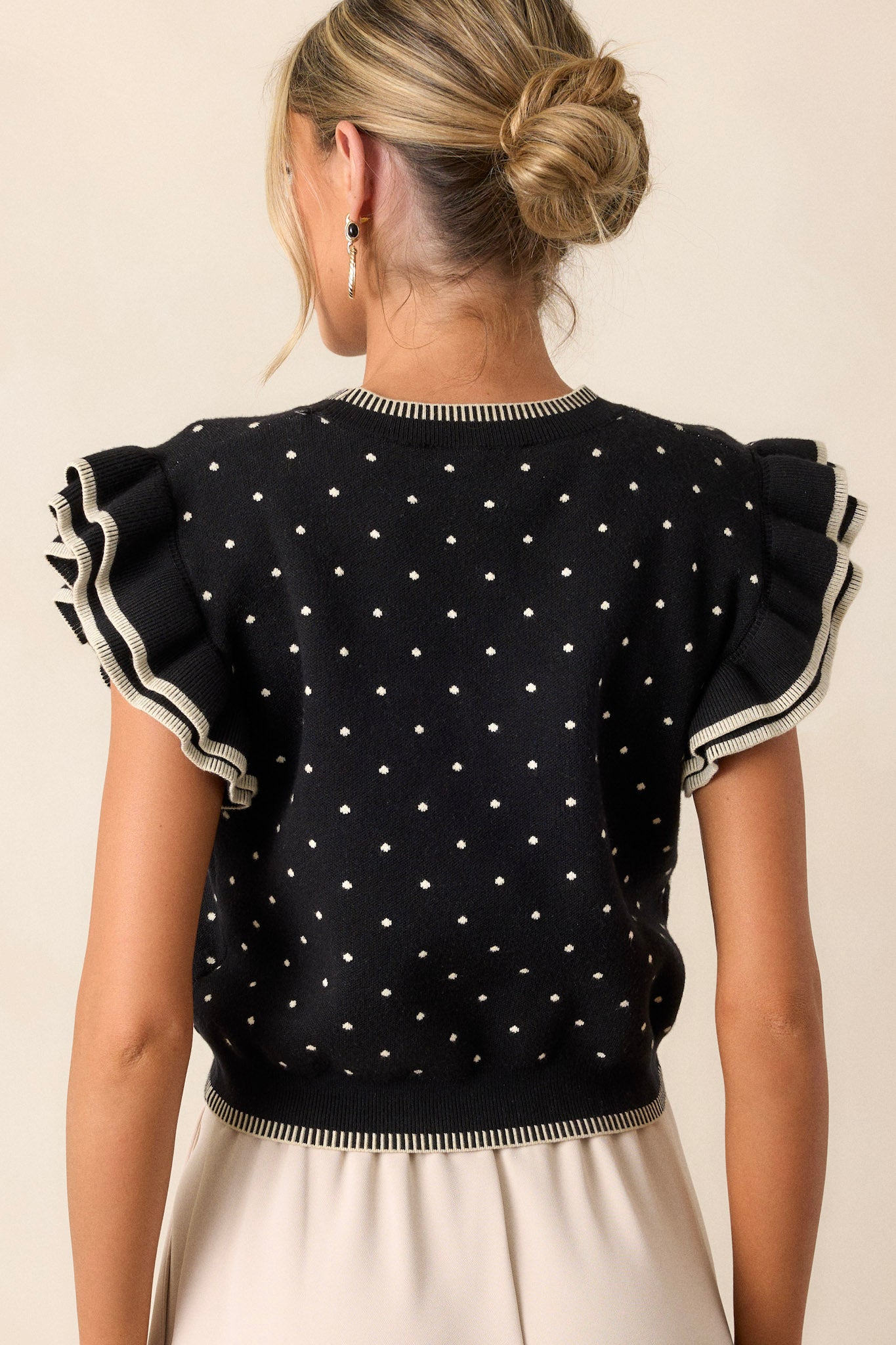 Back view of this black sweater that features an ivory polka dot print, blanket stitching along bottom hem, blanket stitching along the crew neckline, flutter sleeves, and a knit fabric