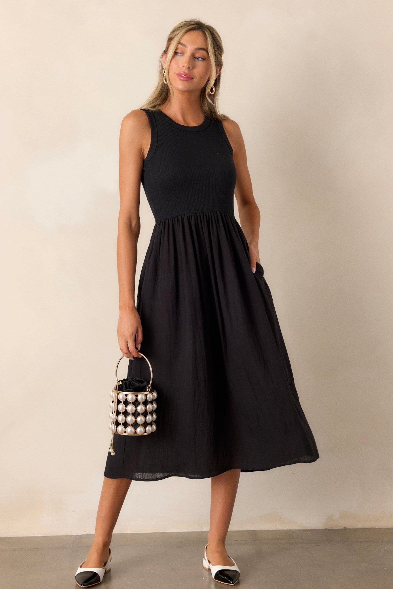 Angled full body view of this sleeveless black dress that features a crew neckline, a stretchy ribbed bust, a cinched waist, and flowing mid-length skirt.