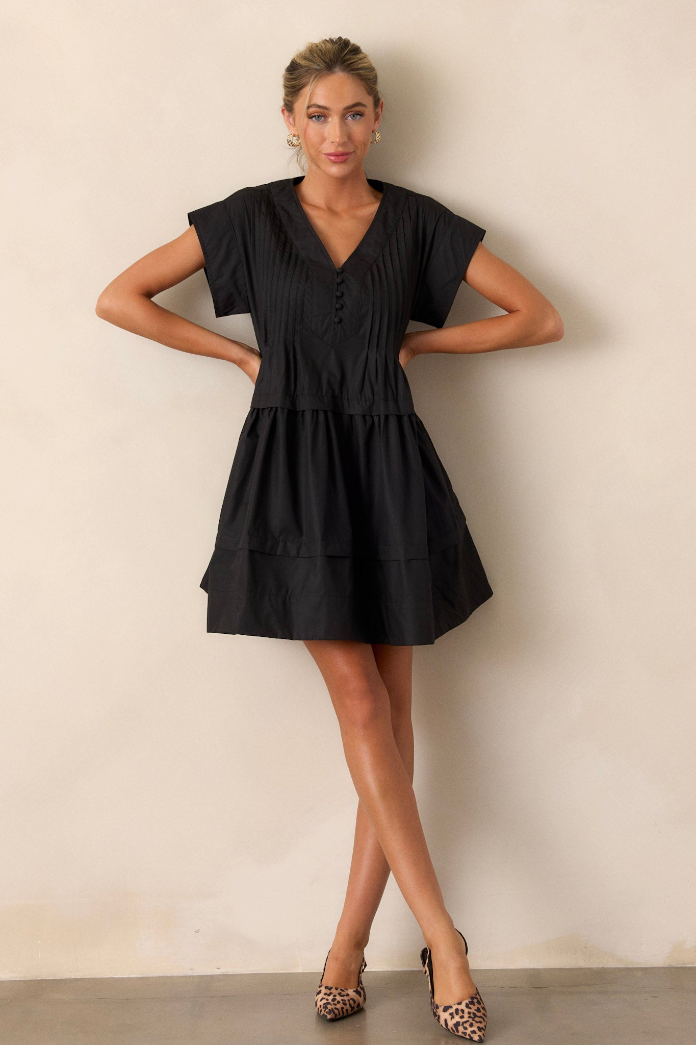 Full length view of the black dress featuring a deep V-neckline, a functional button-front, and two functional side pockets, emphasizing the dress's sleek and versatile style