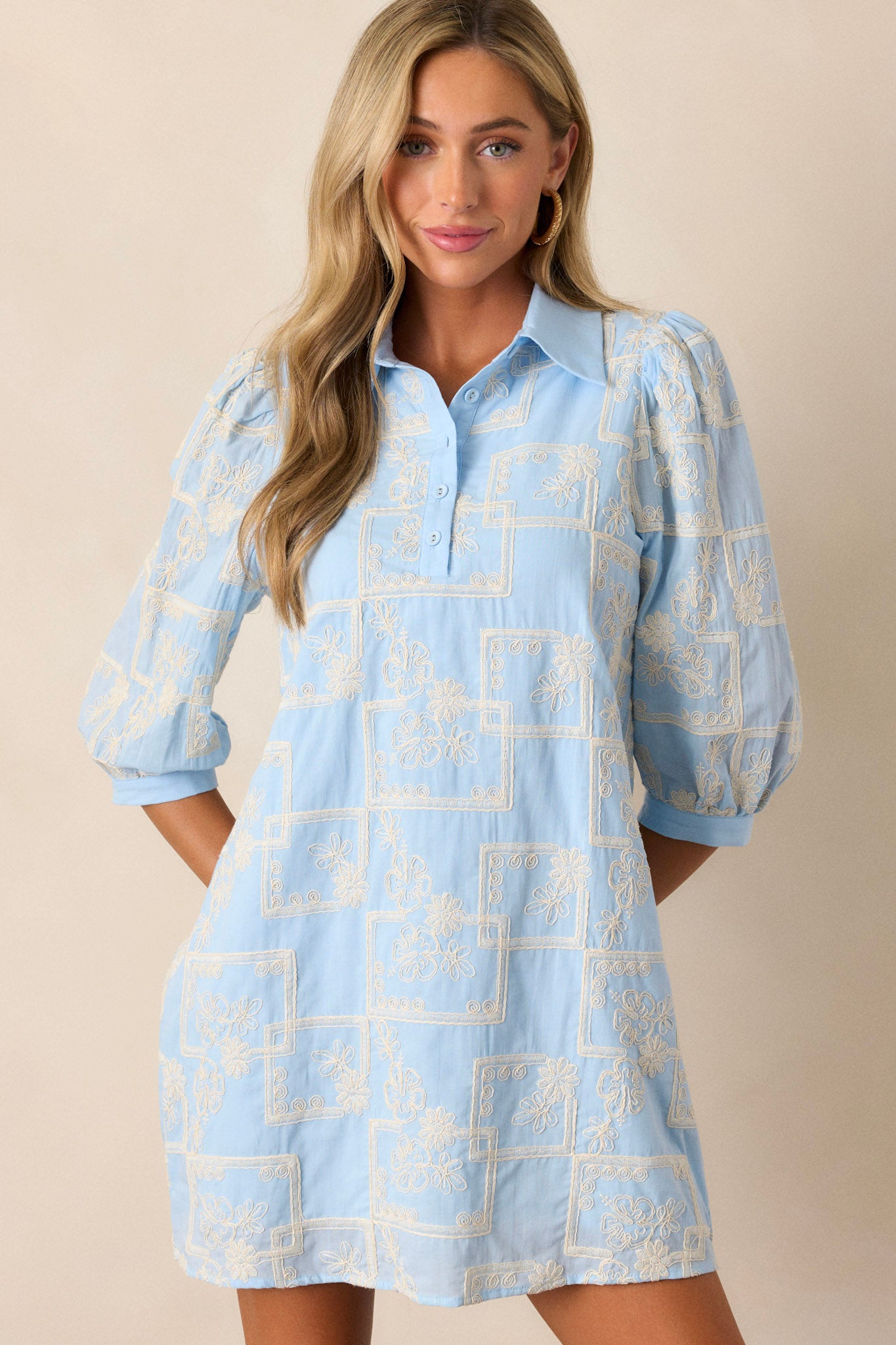 Front view of this dress that features a collared neckline, a functional button up bust, a white embroidered floral and square pattern, a relaxed fit and puffed half sleeves.