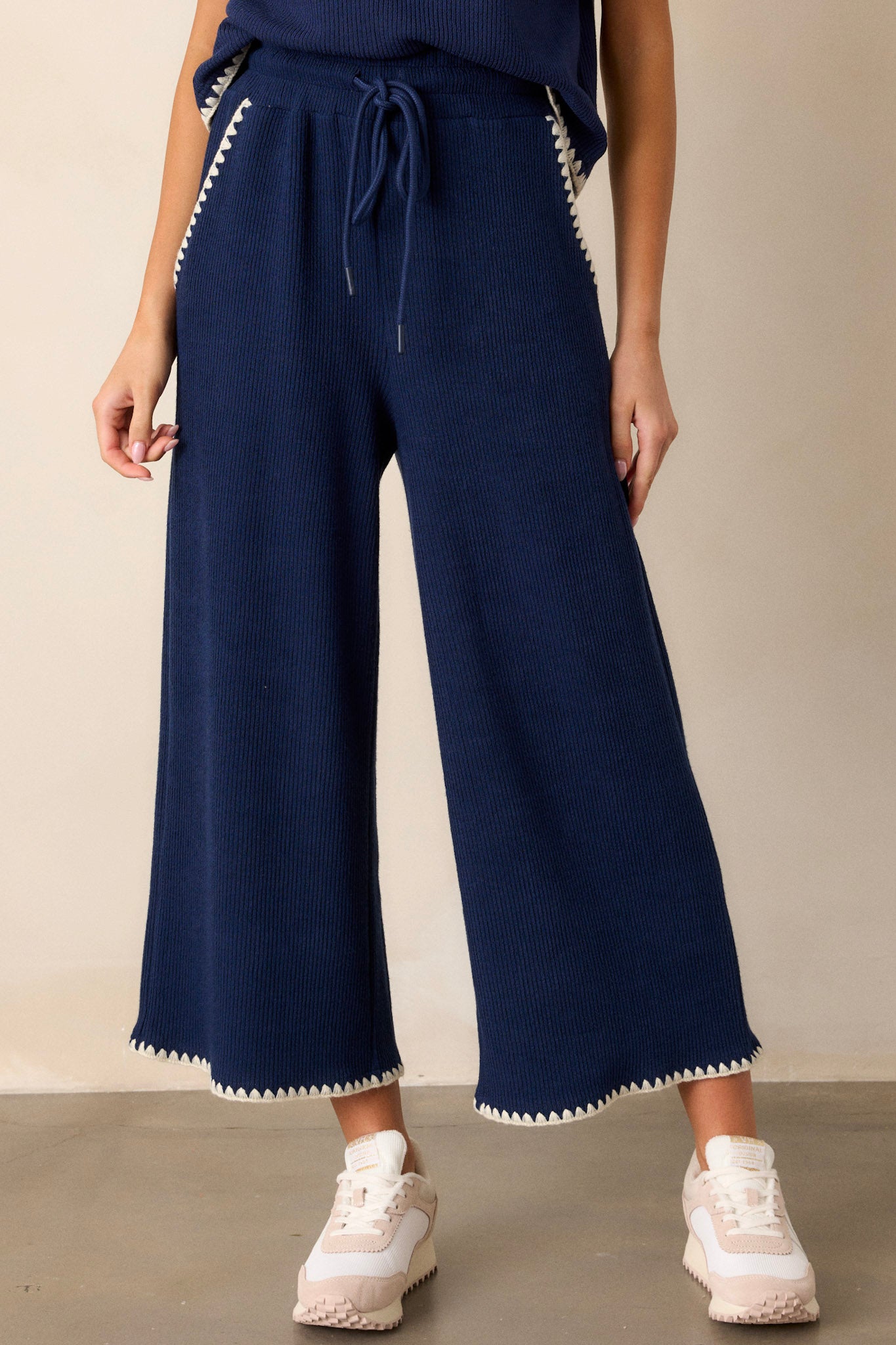 Close-up view of these navy pants that feature an elastic waistband, an adjustable drawstring, ivory blanket stitch detailing, functional hip pockets, a soft knit material, and a wide leg.