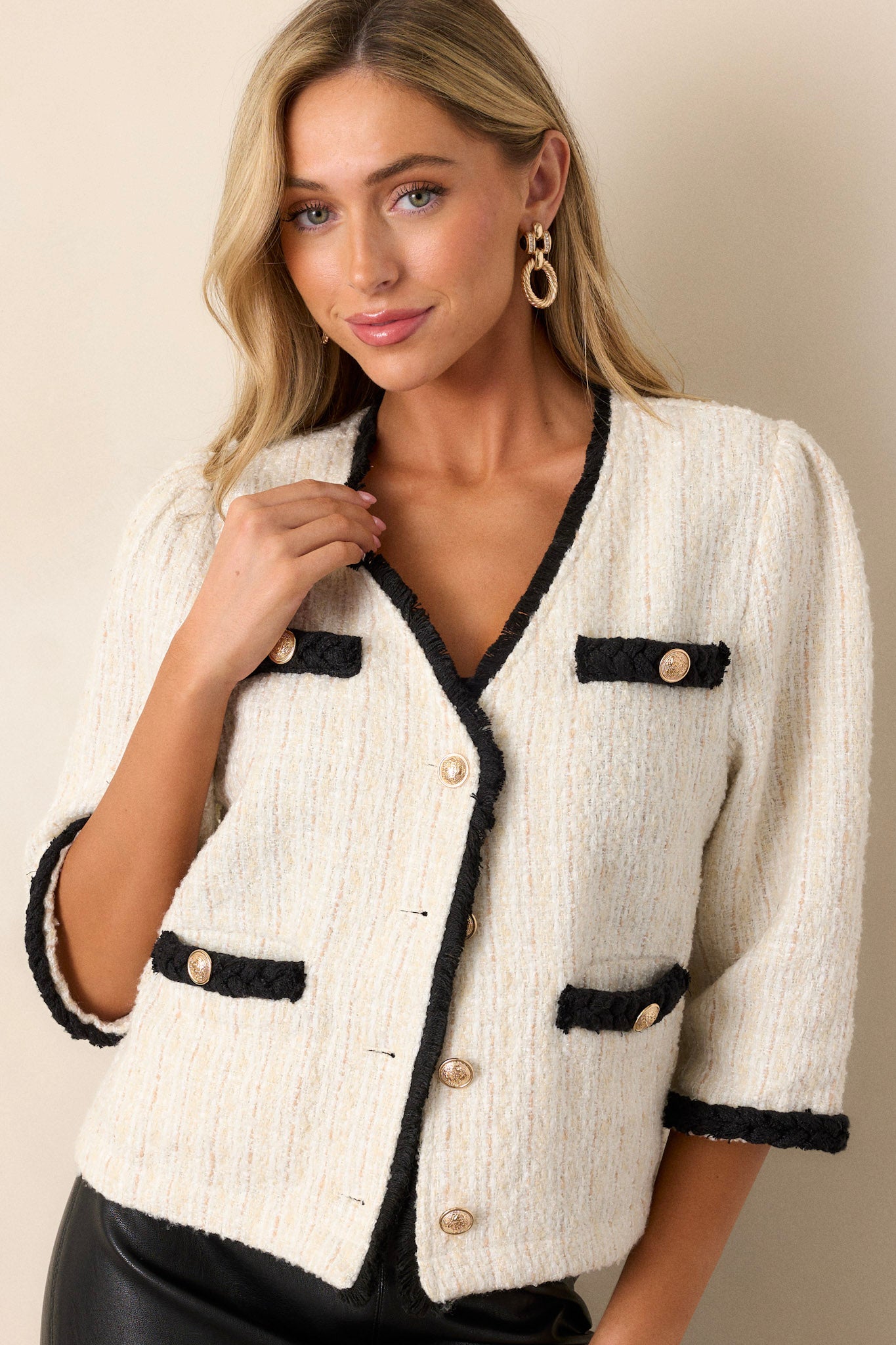 This ivory tweed jacket features a v-neckline, a functional button front, decal chest pockets, functional hip pockets, gold accent buttons and black trim.