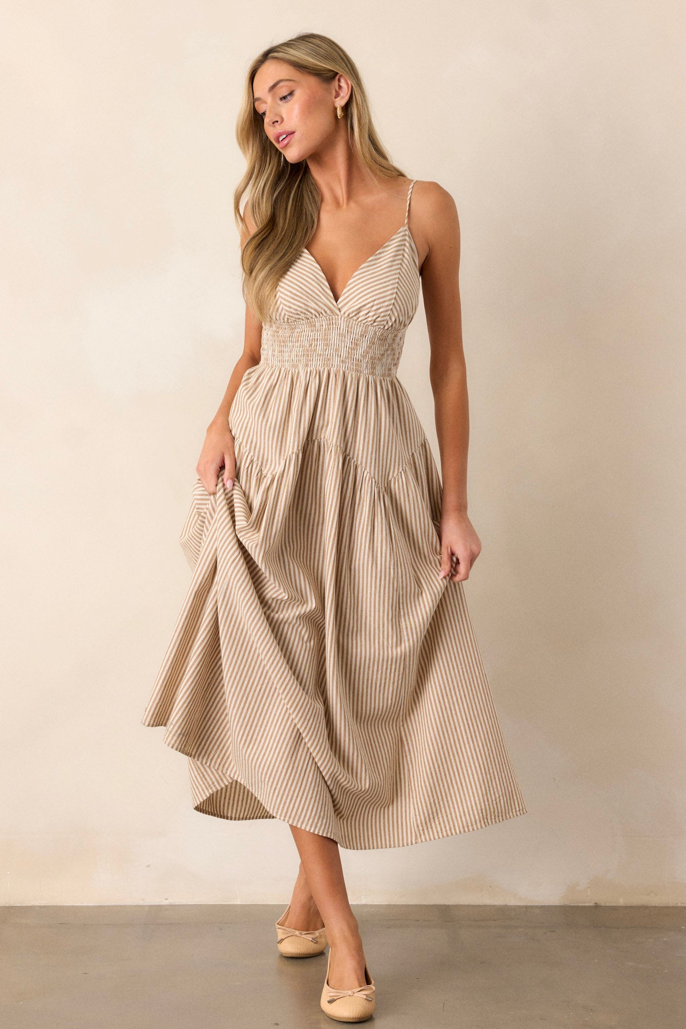 Front view of this tan midi dress  that features a deep v-neckline, a smocked waistband, functional hip pockets, a stripe print, and adjustable spaghetti straps.