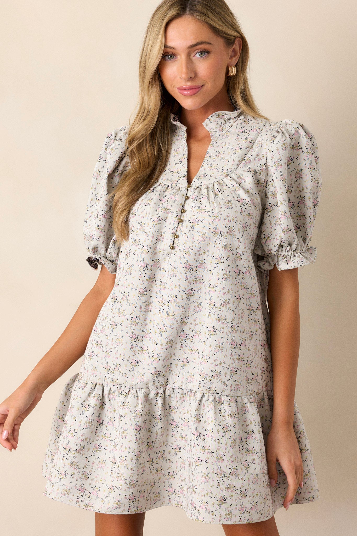 Front angled view of this almond floral dress that features a ruffle collared neckline, a split neckline with a functional button up bust, a soft quilt texture, a small floral pattern, a tiered design, a ruffled trim and short elastic cuffed puff sleeves.