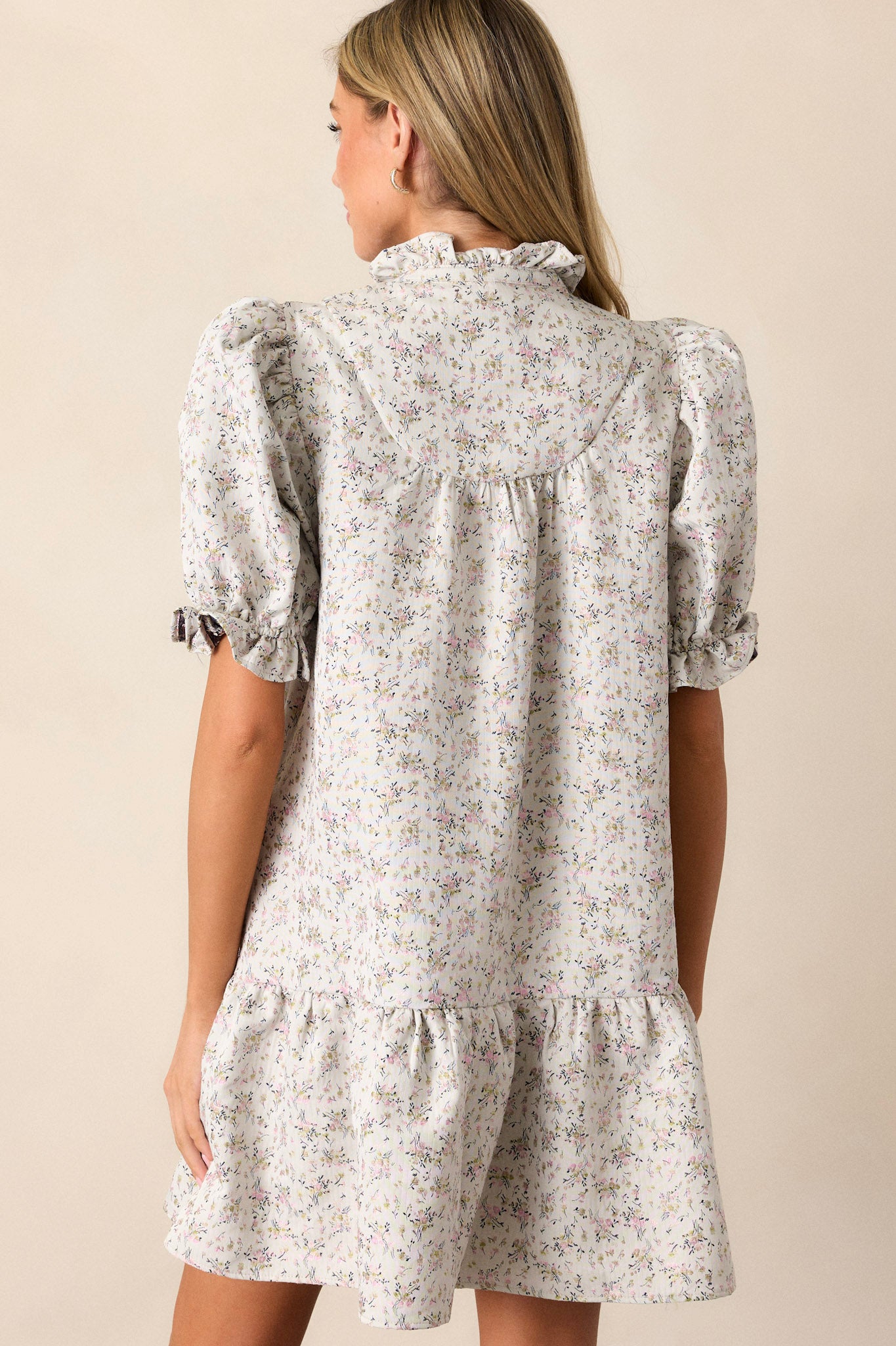 Back view of this almond floral dress that features a ruffle collared neckline, a split neckline with a functional button up bust, a soft quilt texture, a small floral pattern, a tiered design, a ruffled trim and short elastic cuffed puff sleeves.