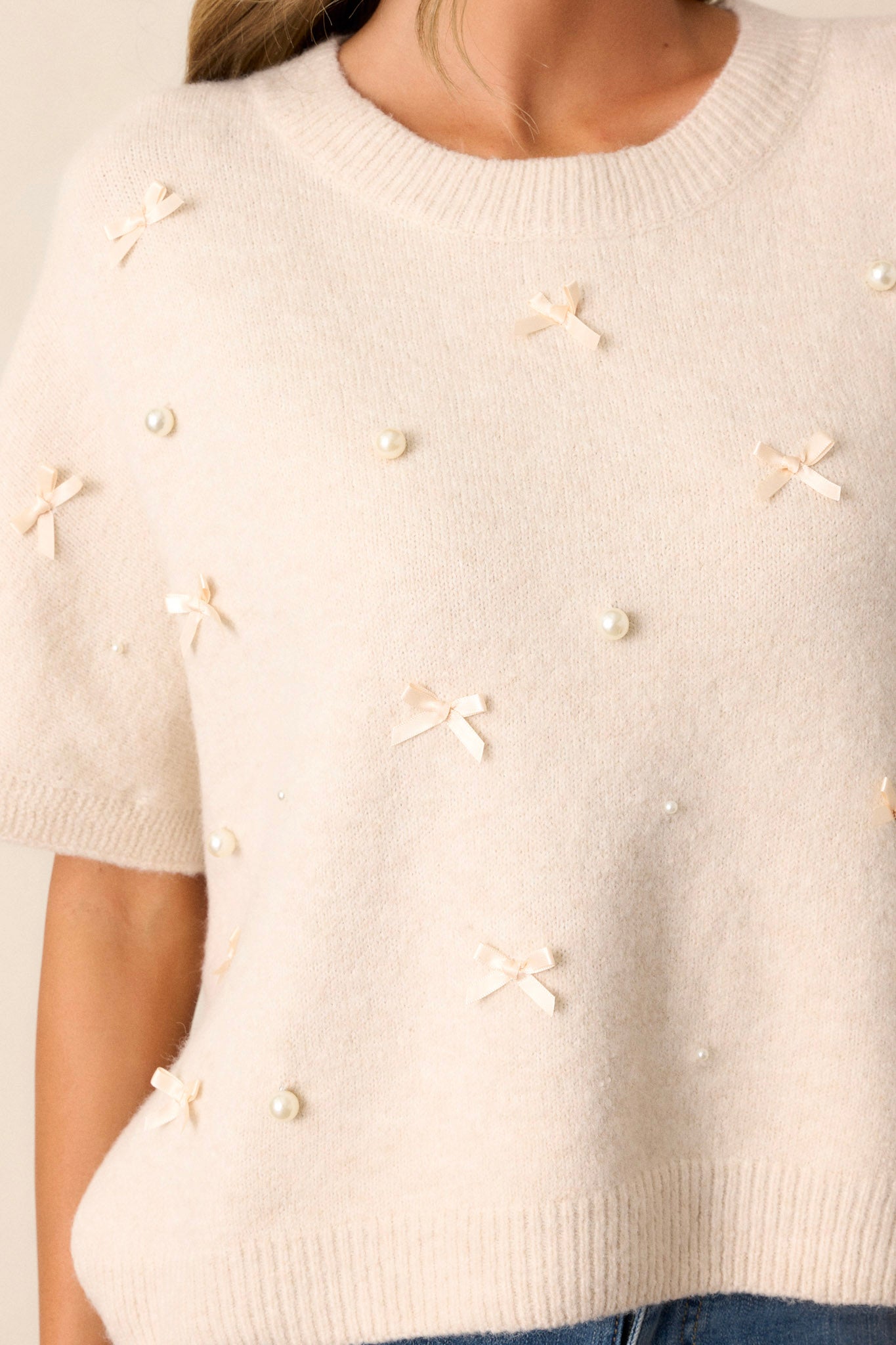 Close-up of this ecru top that features a rounded neckline, an oversized fit, faux pearls and little bows decorating the front, a free cut hem and short sleeves.