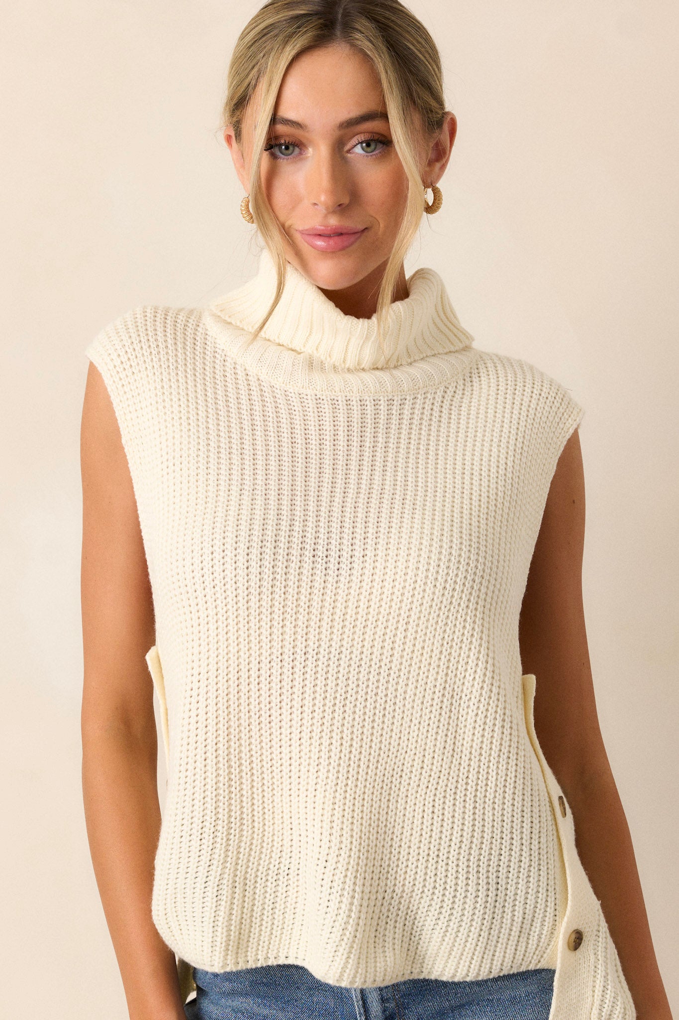 Front view of this ivory sweater that features a turtleneck, a ribbed texture, multiple buttons on side, and a sleeveless design.