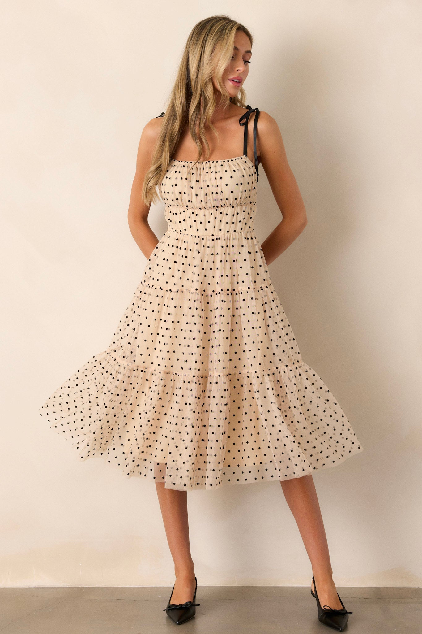 Action shot of a beige midi dress displaying the fit and movement, highlighting the square neckline, thin self-tie straps, elastic waistline, tiers, black polka dots, and tulle.