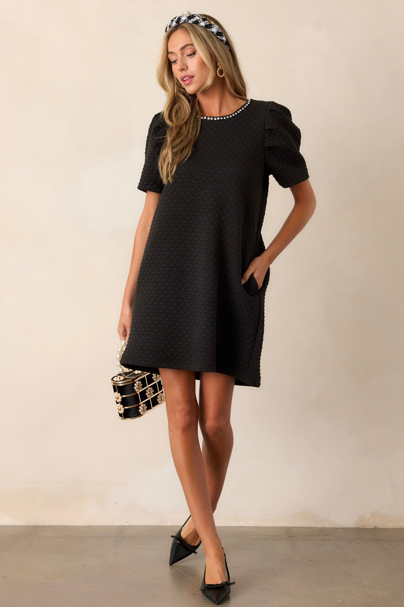 Full body view of this black dress featuring a crew neckline, rhinestone embellishments around neckline, puff sleeves, a textured material, and a back cutout with loop closure.