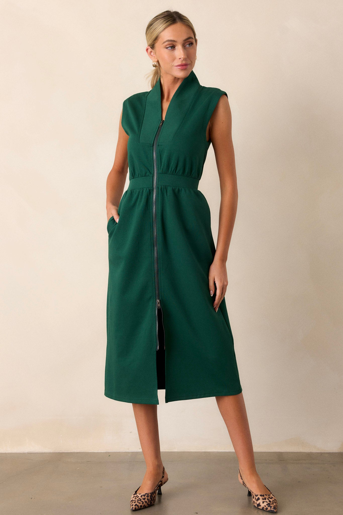 Angled view of this hunter green dress that features a v-neckline, a slight collar, a zipper front, functional hip pockets, a middle split in the hem and a sleeveless design.