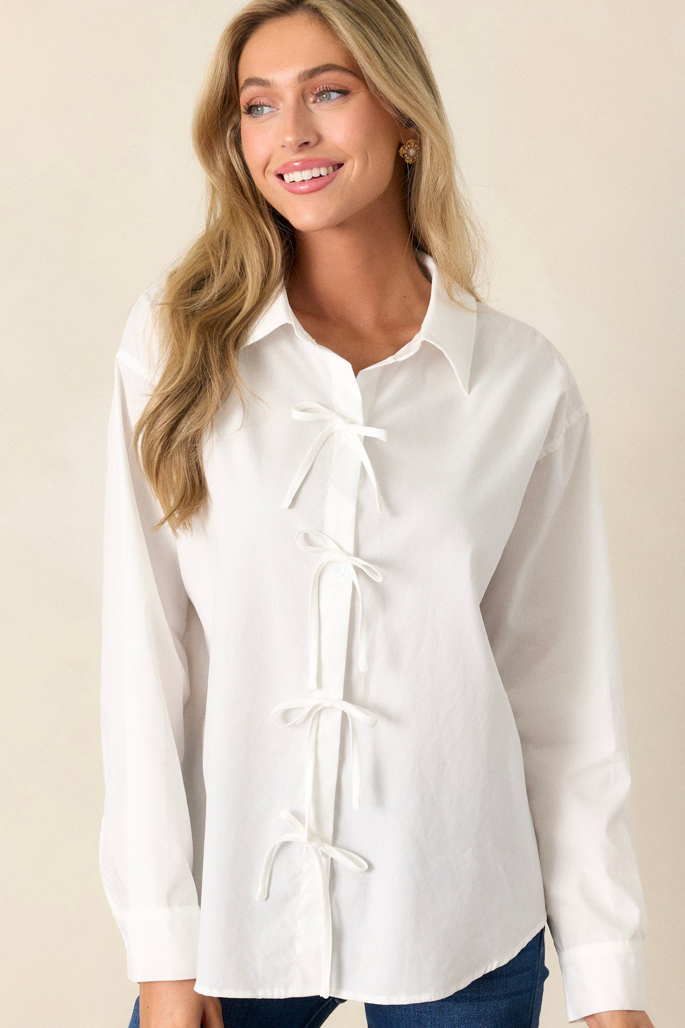 Front angled view of a white button-front top featuring a collared neckline, functional button front, white bow details, a relaxed fit, and long sleeves