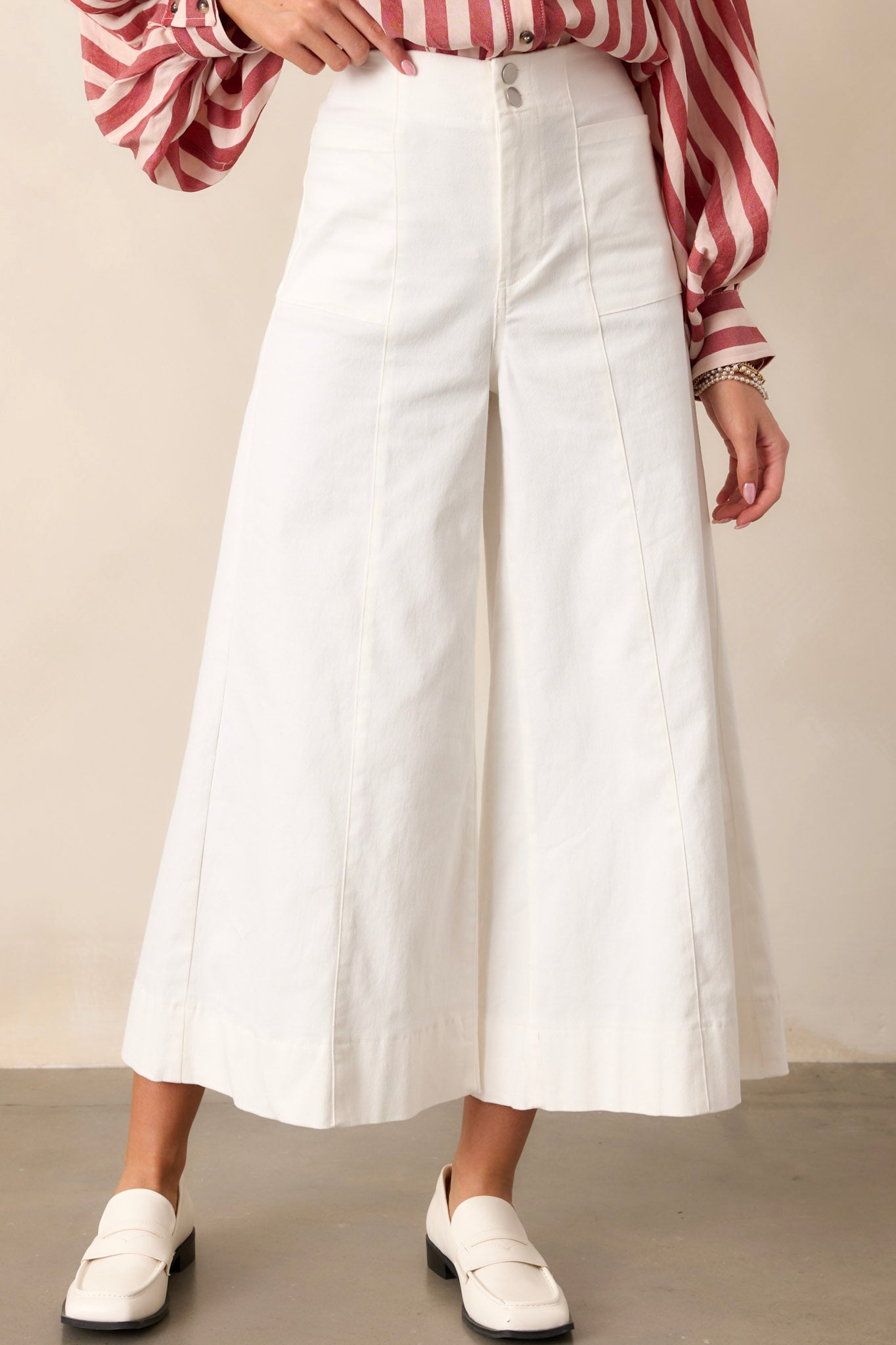 Front angled view of white pants featuring a high waisted design, a double button closure with a zipper, functional front pockets, front & back seams, and a wide leg with a cropped length