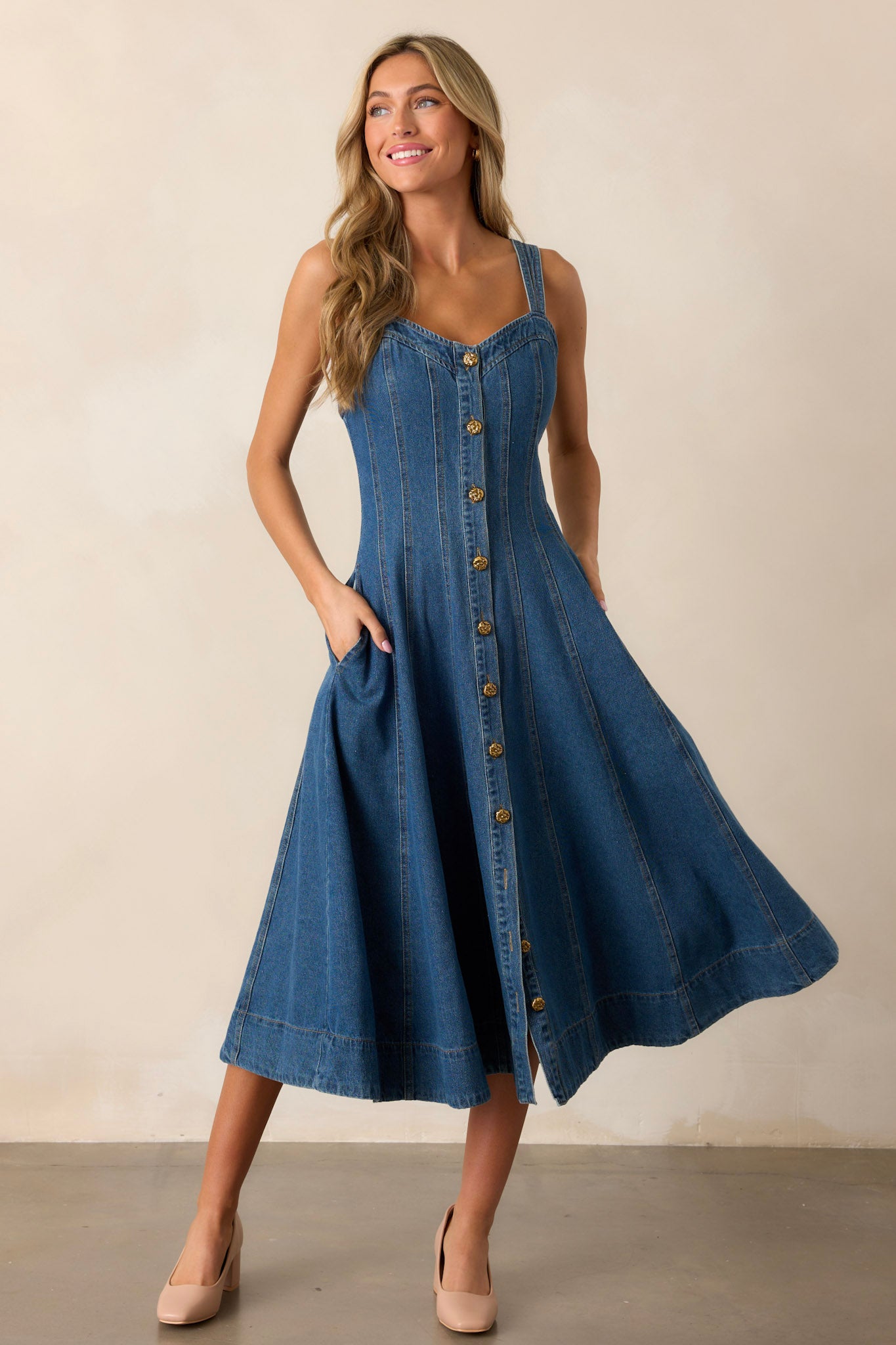 Front view showing the movement of this dark wash midi dress made with high-quality denim, featuring a sweetheart neckline, thin straps, functional pockets, functional buttons down the length of the dress, and vertical seam detailing.