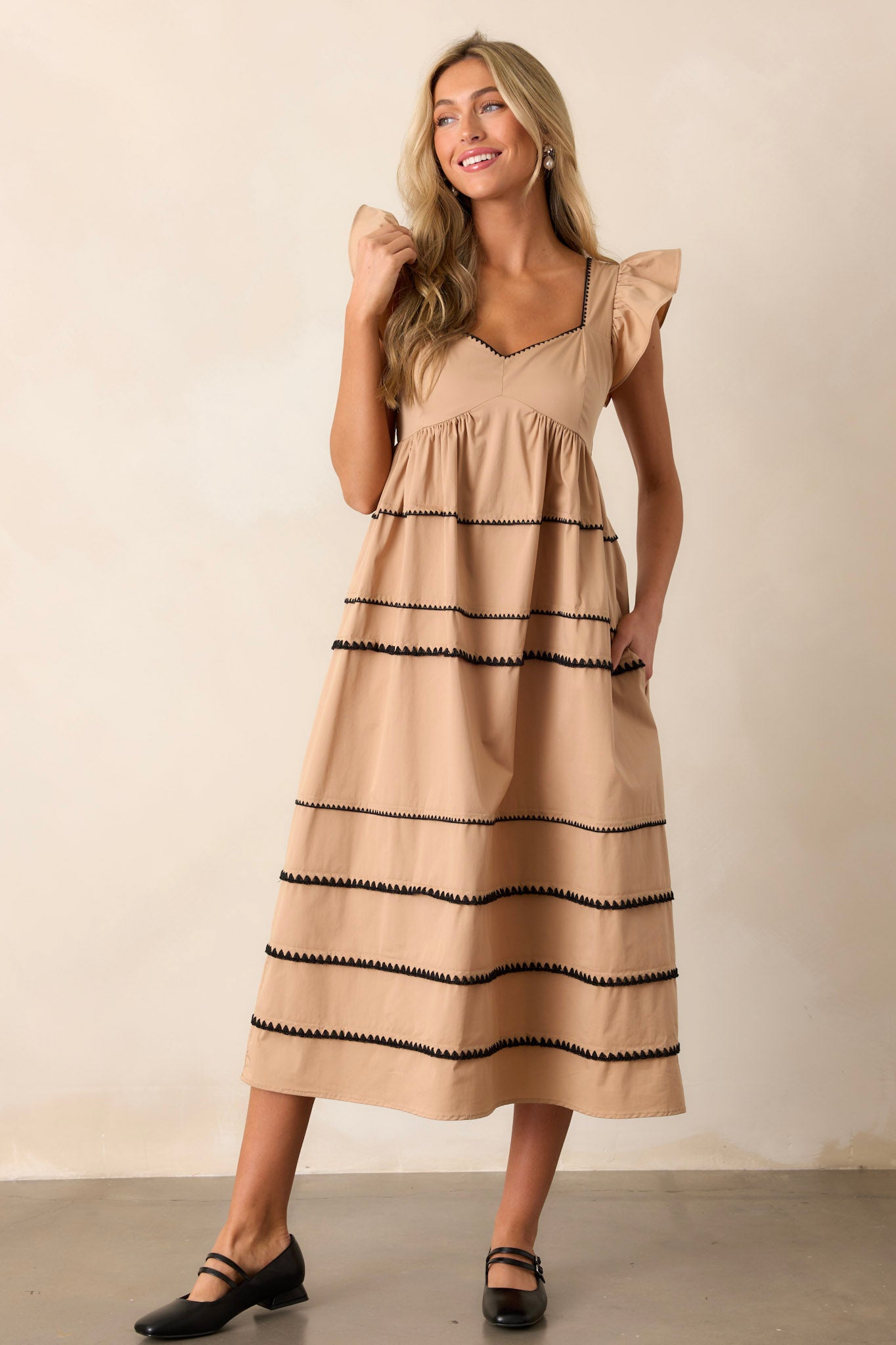 Front angled view of the tan dress featuring ruffle cap sleeves, a smocked back, multiple tiers, two functional pockets, and ric-rac detailing