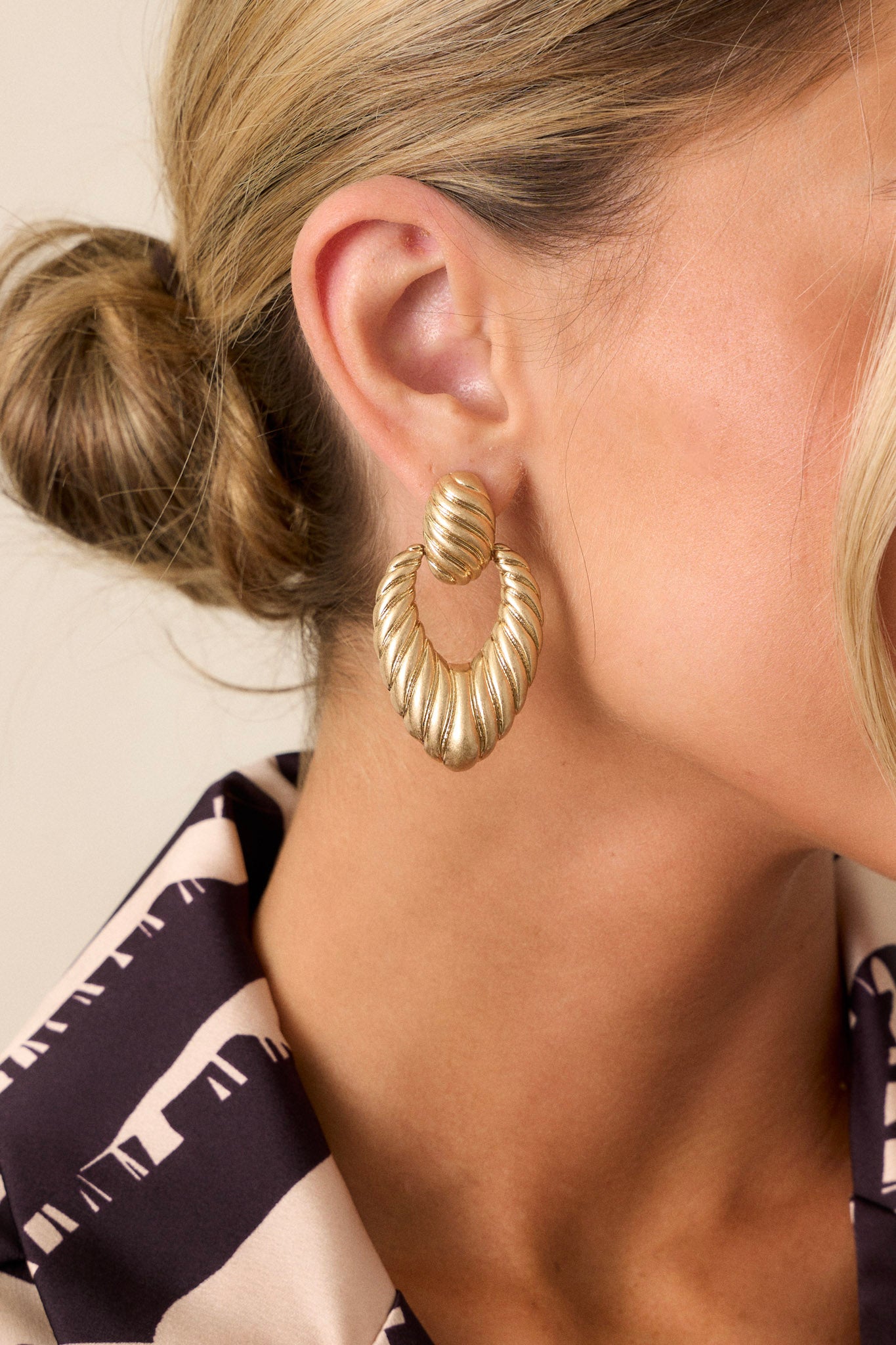 A close-up of gold earrings featuring a textured oval stud connected to a textured triangular hoop, with secure post backings.