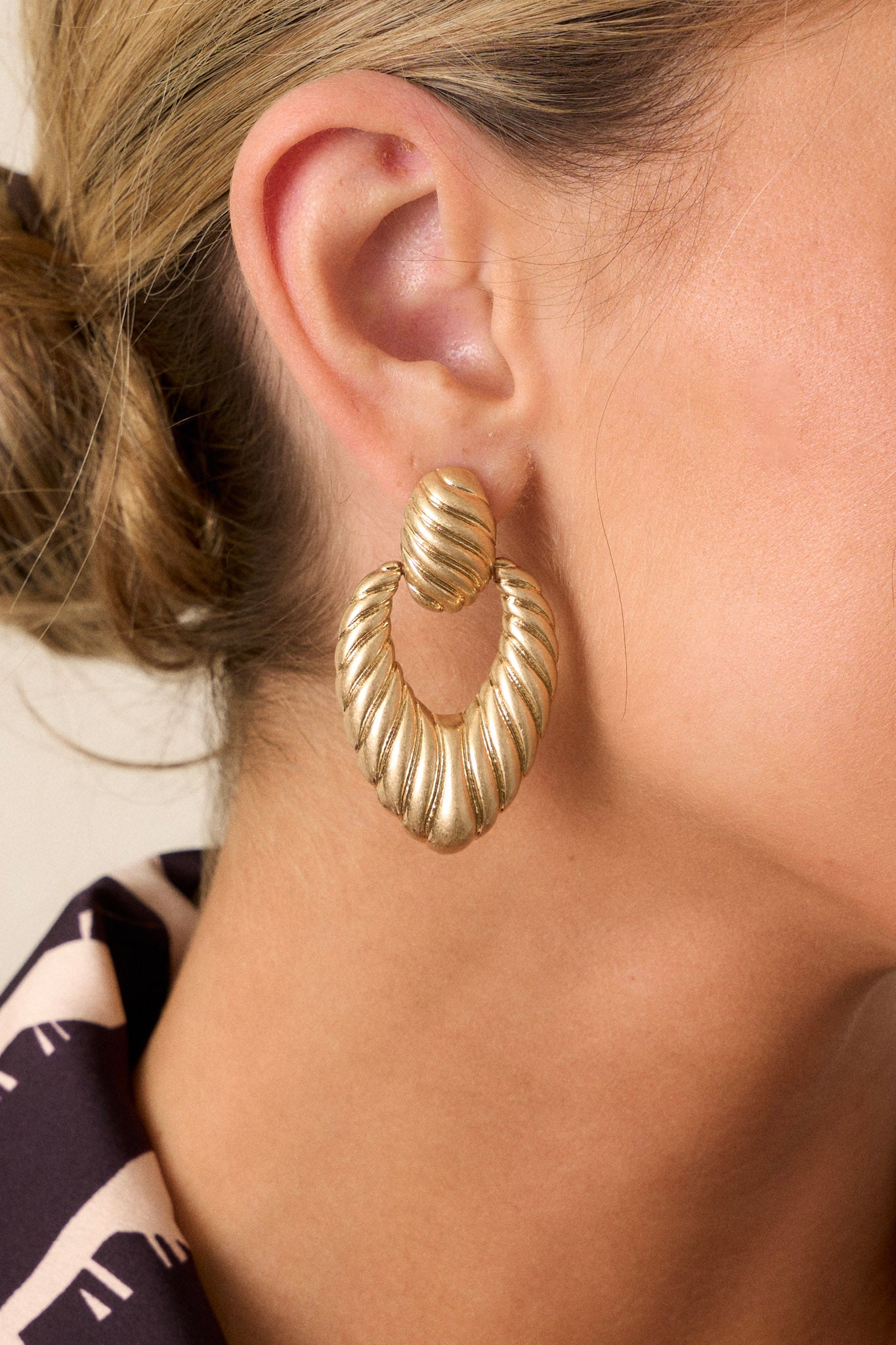 Extreme close-up of gold earrings highlighting the intricate texture of the oval stud and triangular hoop, with visible secure post backings.