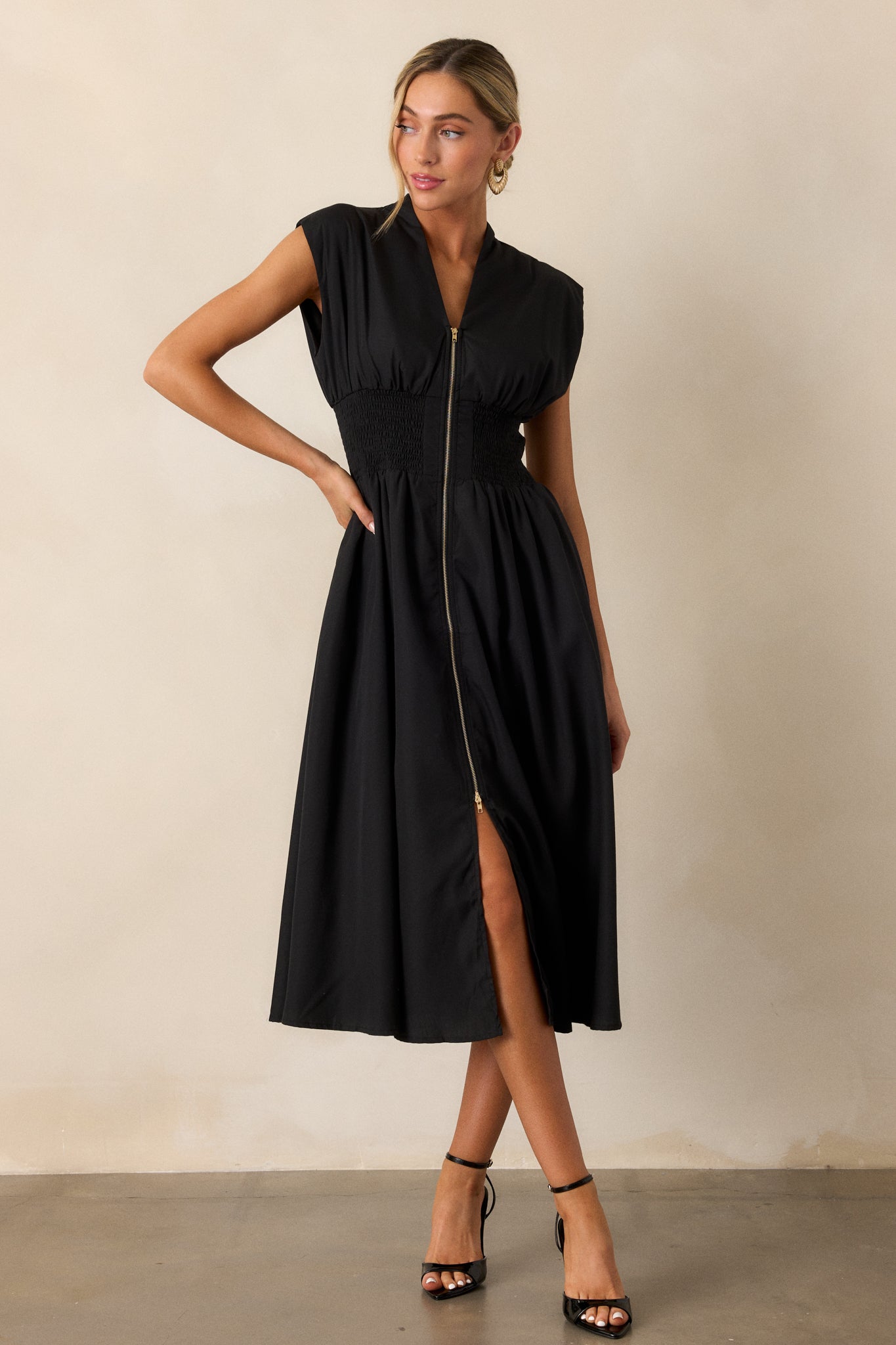 This black midi dress features a v-neckline, padded shoulders, a full zipper front, a fully smocked waist, functional hip pockets, and a front slit.