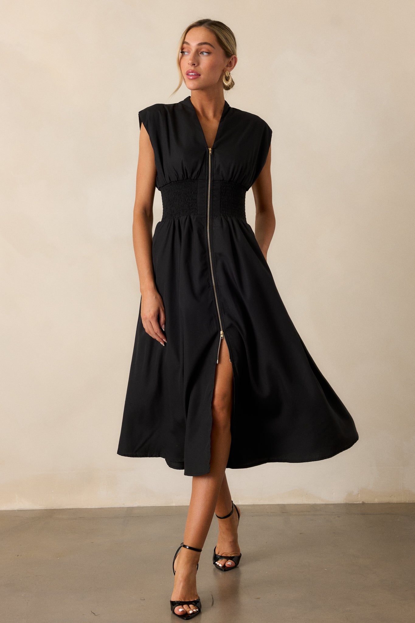 Front angled view of a black midi dress featuring a v-neckline, padded shoulders, a full zipper front, a fully smocked waist, functional hip pockets, and a front slit