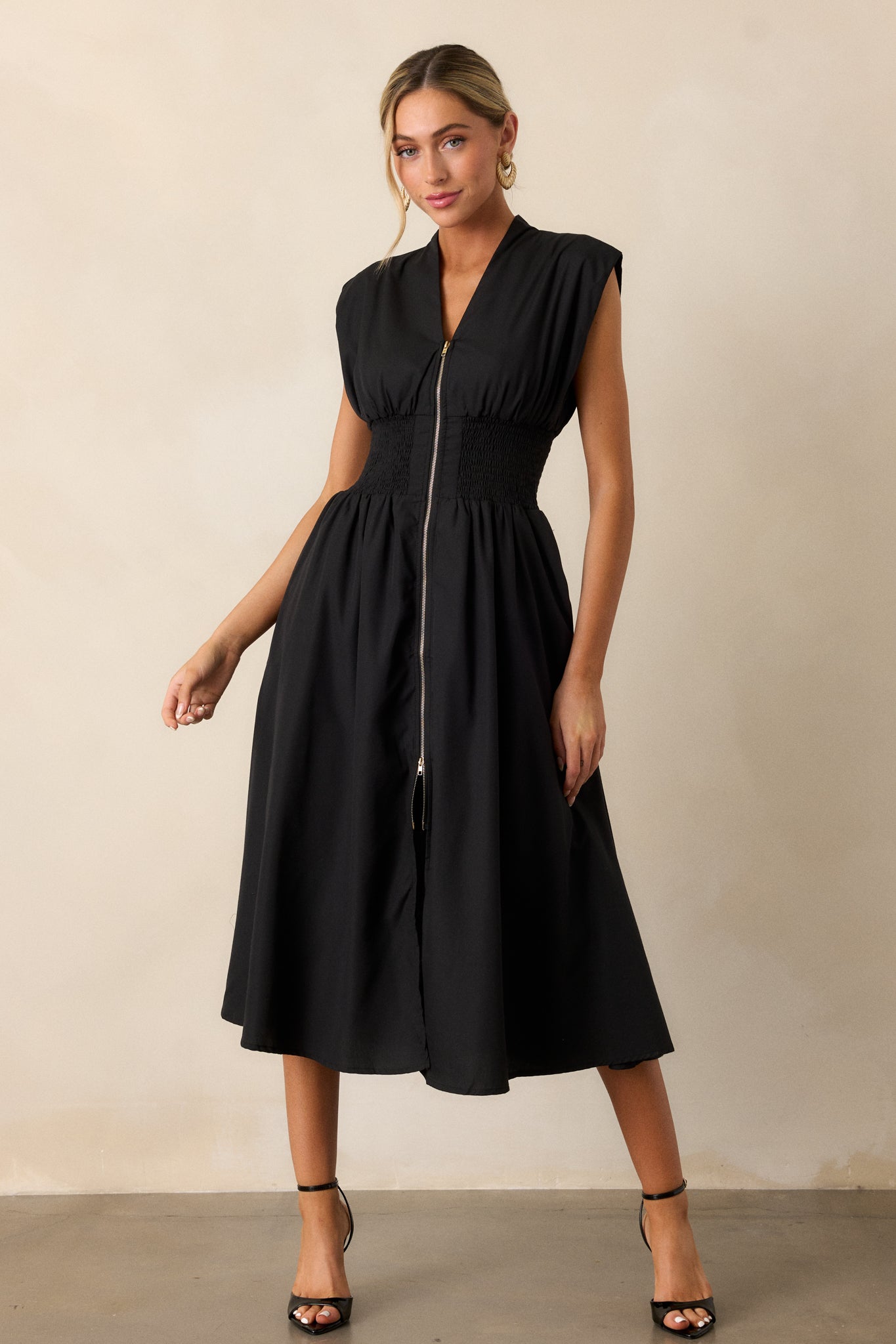 Full length view of a black midi dress with a v-neckline, padded shoulders, a full zipper front, a fully smocked waist, functional hip pockets, and a front slit