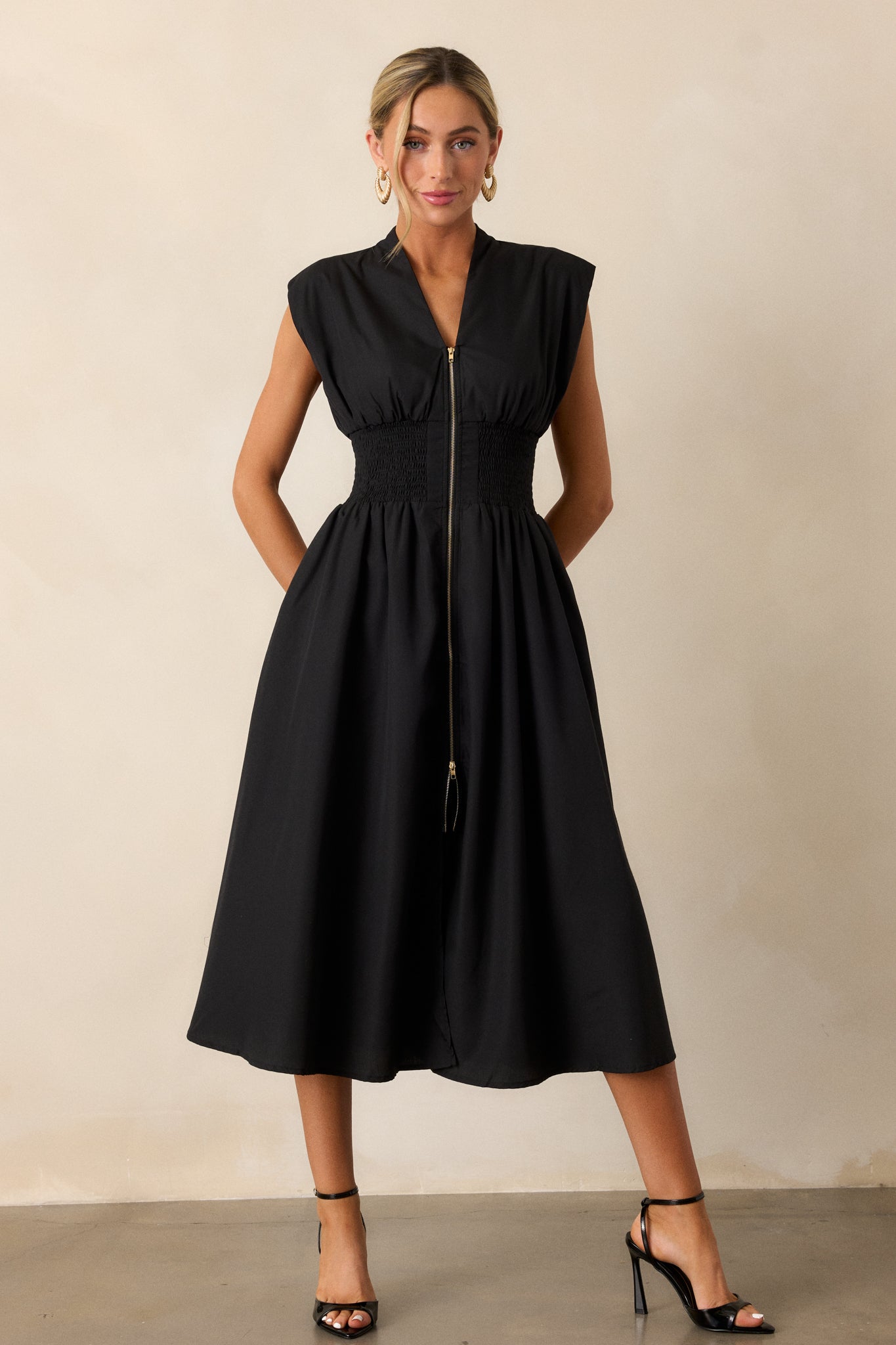 Action shot of a black midi dress displaying the fit and movement, highlighting the v-neckline, padded shoulders, full zipper front, fully smocked waist, functional hip pockets, and front slit.