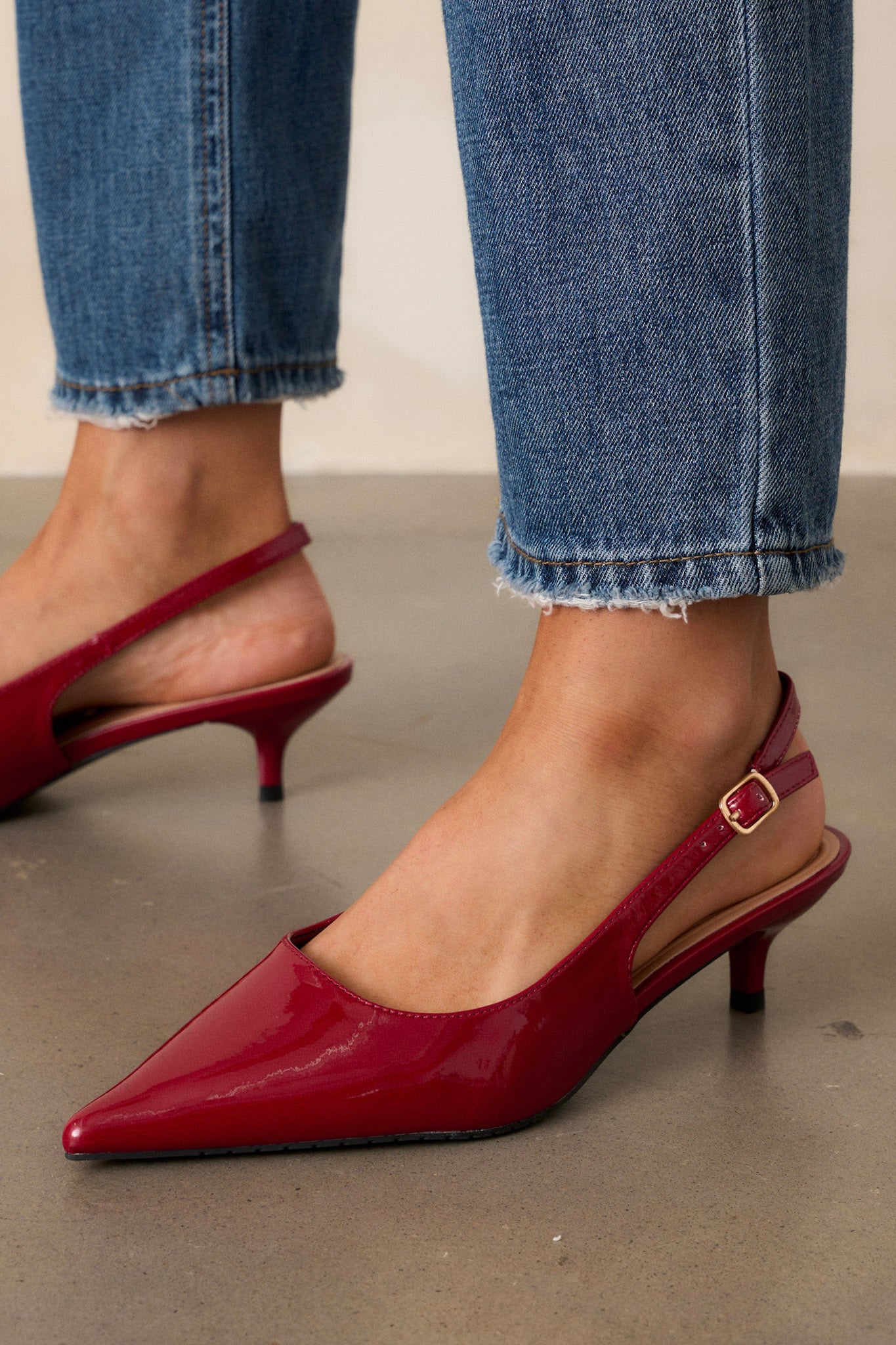 Close-up of a model wearing burgundy heels with a pointed toe and glossy patent finish, featuring an adjustable slingback strap and a gold buckle.