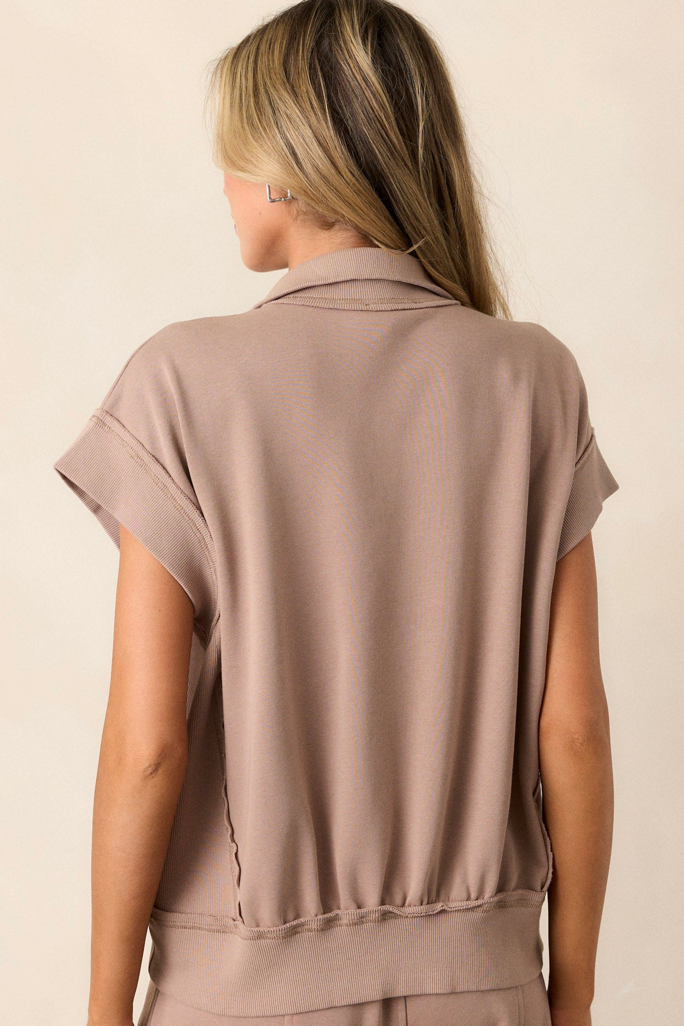 Back view of this light mocha top that features a collared neckline, a functional zipper bust, a functional kangaroo pocket, a thick ribbed hemline, contrasting texture on the sides and shirt sleeves.