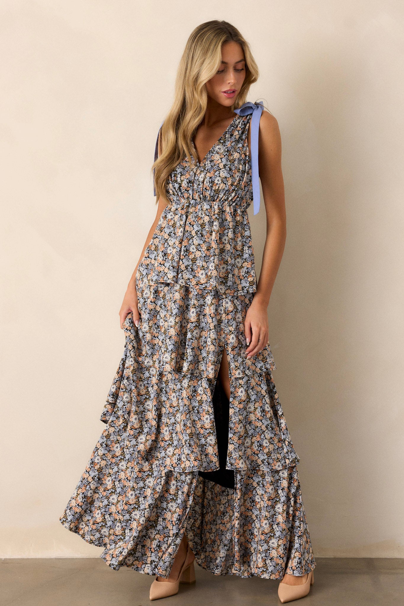 Angled full body view of this black floral maxi dress that features a v-neckline, self-tie shoulder features, an elastic waistline, multiple tiers, a side slit, and a flowing silhouette.