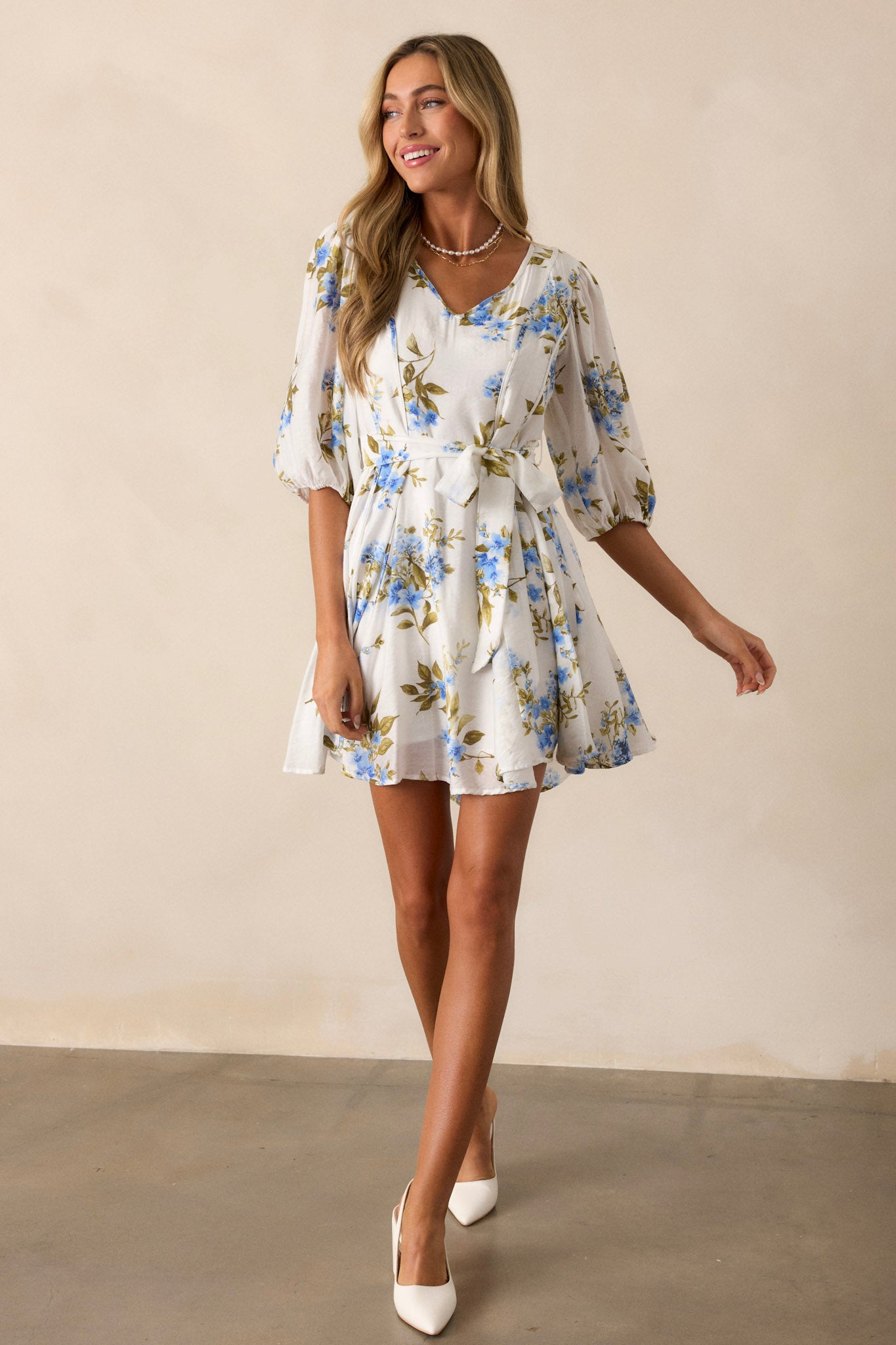 Full body photo of this blue floral dress with puff sleeves, featuring a V-neckline and a tied waist, styled with white wedge sandals.