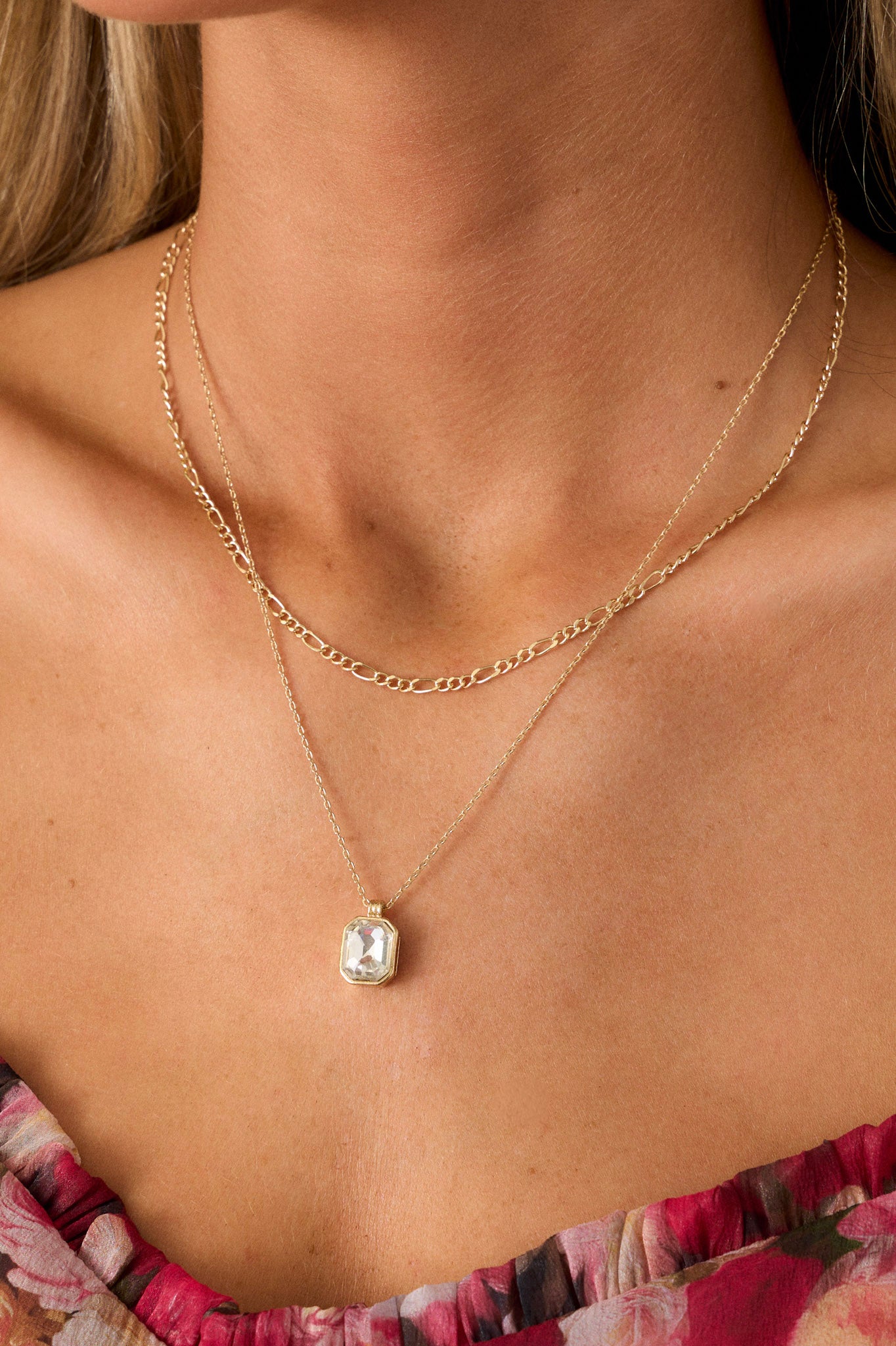 Close up view of this gold necklace featuring a shorter figaro chain, and a longer cable chain with a rectangle bezel-set rhinestone pendant.