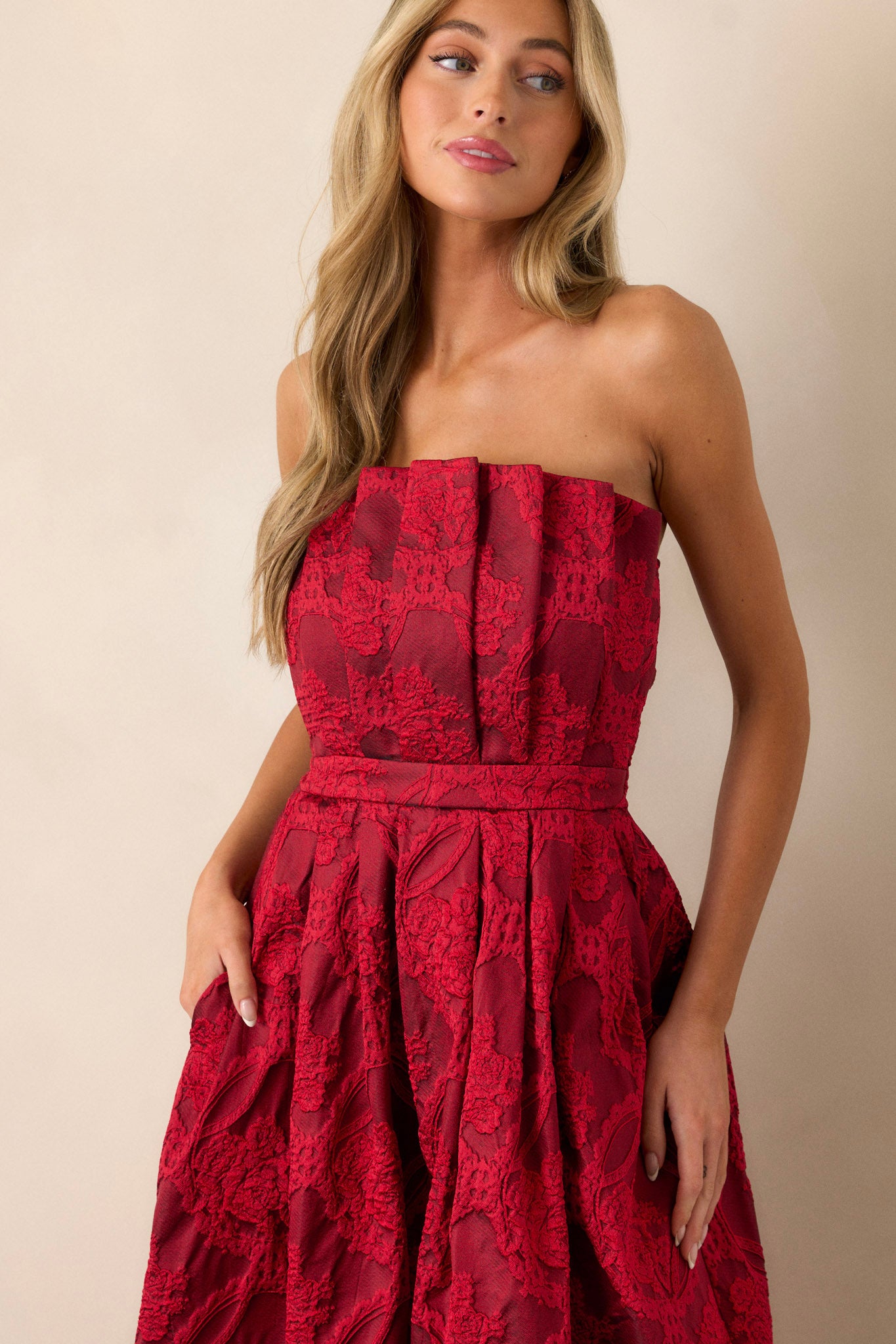 Front view of a red midi dress featuring a pleated bodice, a slightly pleated skirt, a jacquard design, functional hip pockets, a smocked back, a discrete back zipper and a strapless design.