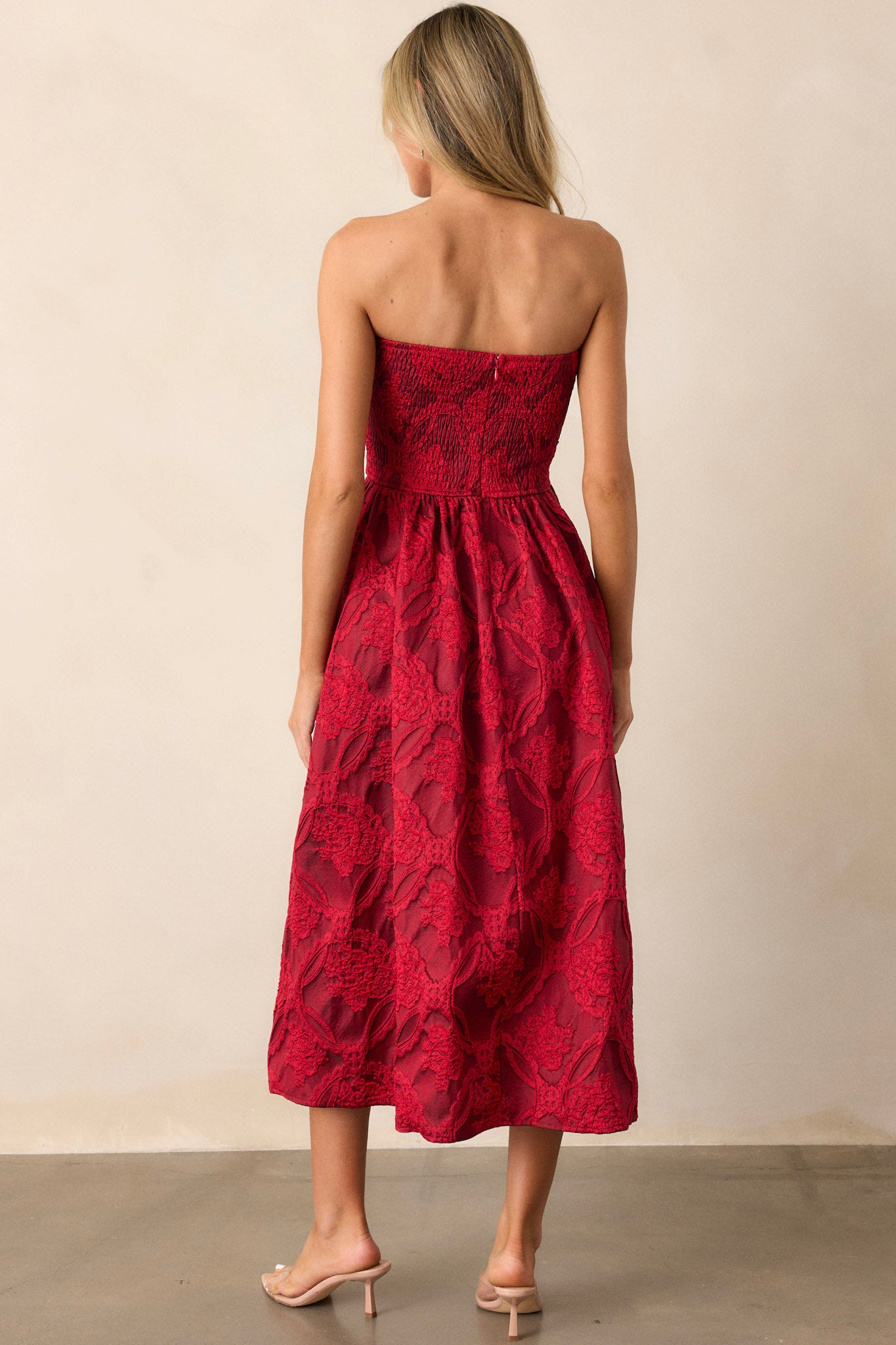 Back view of a red midi dress featuring a pleated bodice, a slightly pleated skirt, a jacquard design, functional hip pockets, a smocked back, a discrete back zipper and a strapless design.