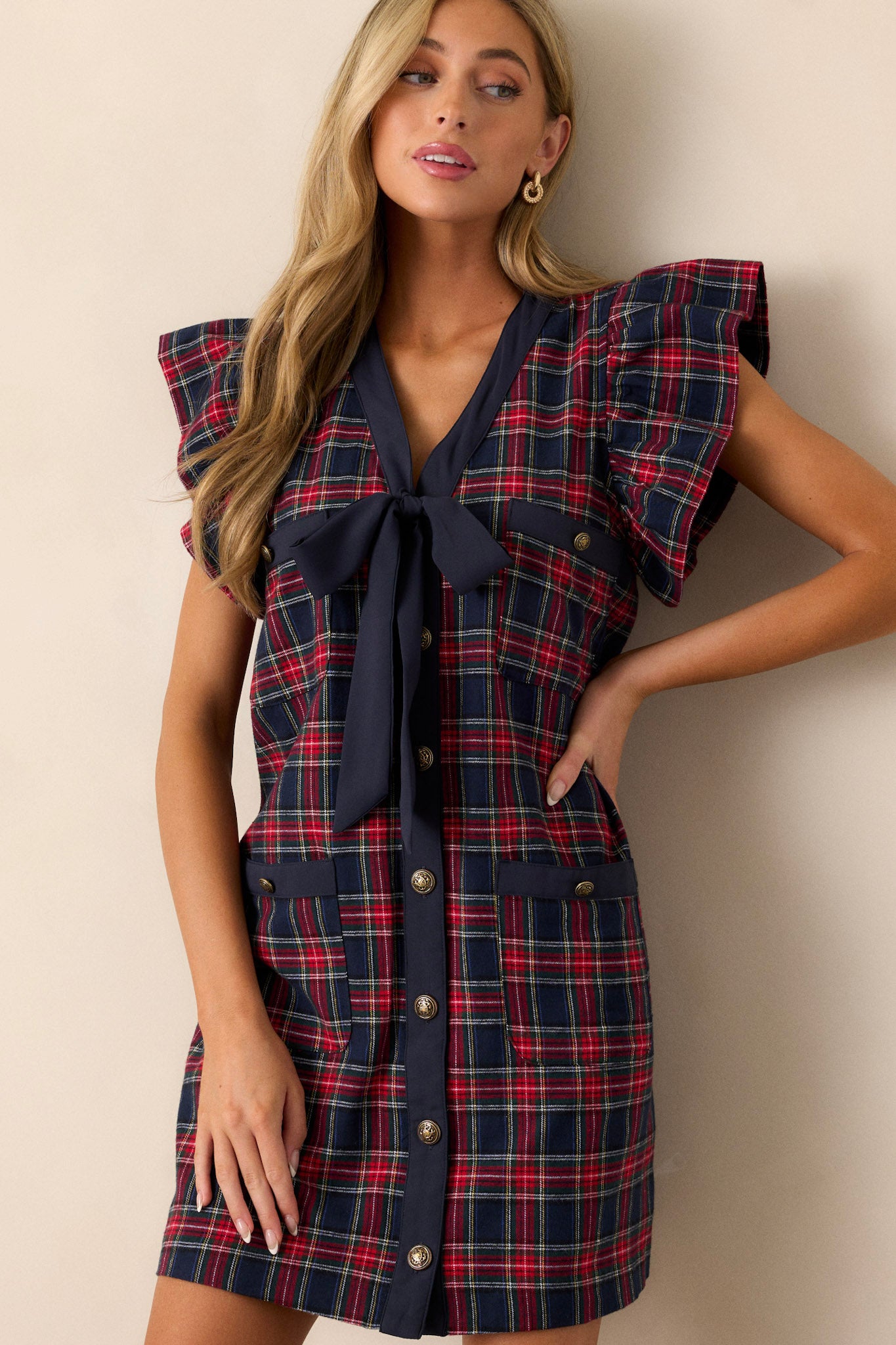 This navy mini dress features a classic plaid print, a navy bow detail on the v-neckline, functional antique gold buttons up the front, two sets of functional pockets, flutter sleeves, and a relaxed fit.