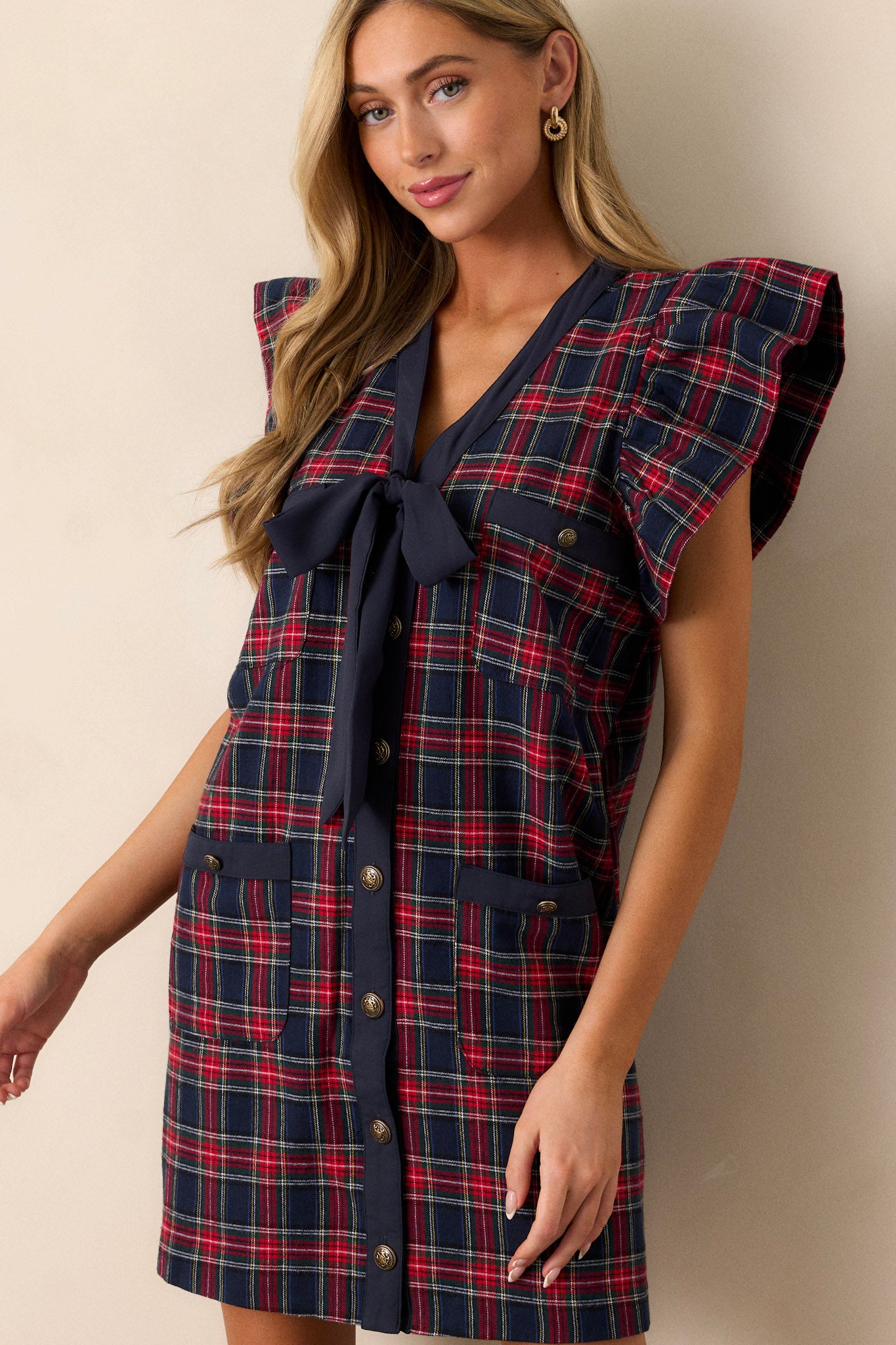 Front view of a navy and red mini dress featuring a classic plaid print, a navy bow detail on the v-neckline, functional antique gold buttons up the front, two sets of functional pockets, flutter sleeves, and a relaxed fit.