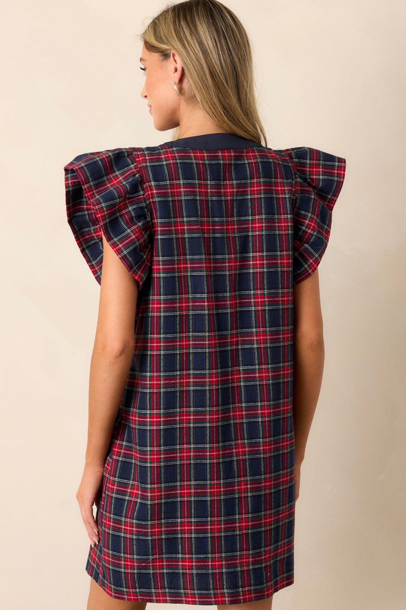 Back view of a navy and red mini dress featuring a classic plaid print, a navy bow detail on the v-neckline, functional antique gold buttons up the front, two sets of functional pockets, flutter sleeves, and a relaxed fit.