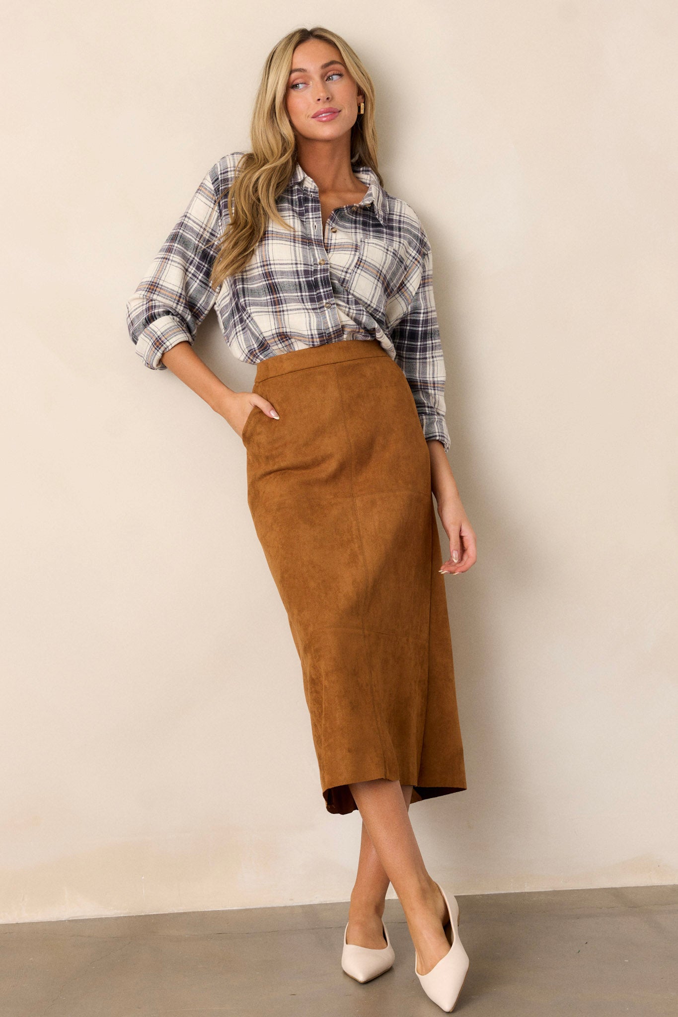 Full body view of a midi skirt featuring a caramel brown faux suede material, an A-line silhouette, functional pockets, and a small slit up the back of the skirt.