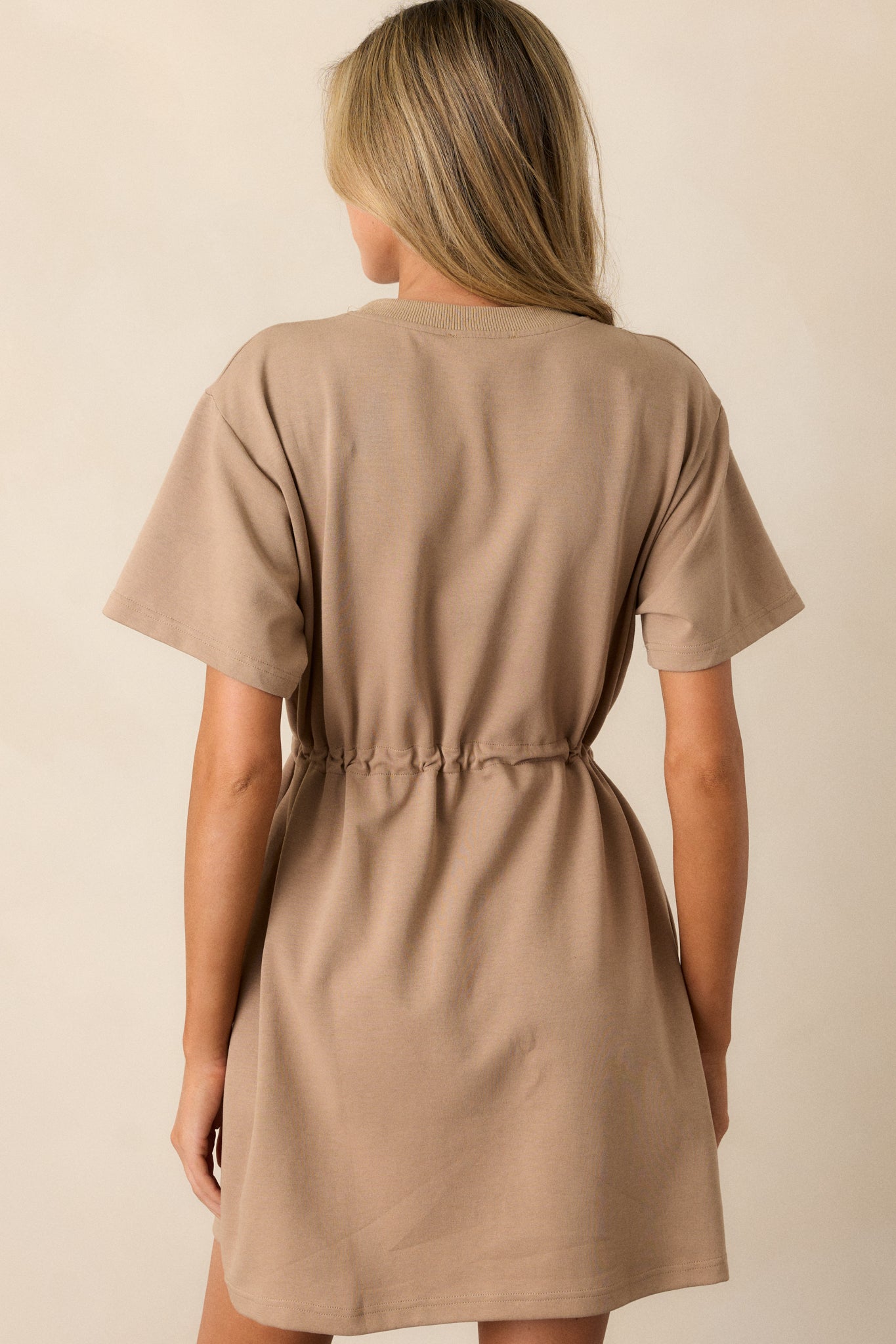 Back view of a light mocha mini dress featuring a crew neckline, a drawstring waist with gold heart shaped charms on the strings, gold aglets, two functional hip pockets, and short sleeves.